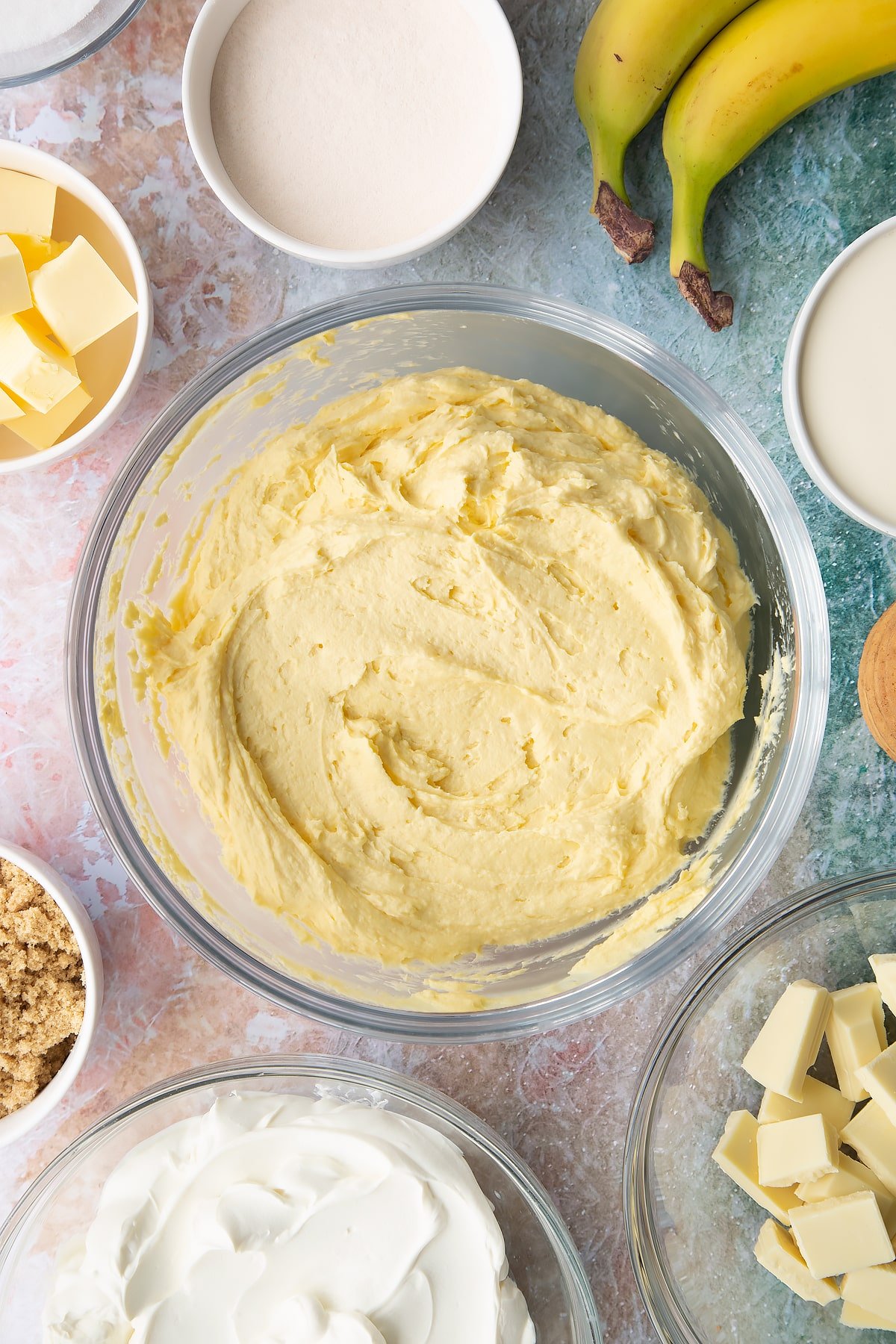 Cream, cream cheese, sugar, banana pudding powder and melted white chocolate whisked together in a bowl. Ingredients to make banoffee pie cheesecake surround the bowl.