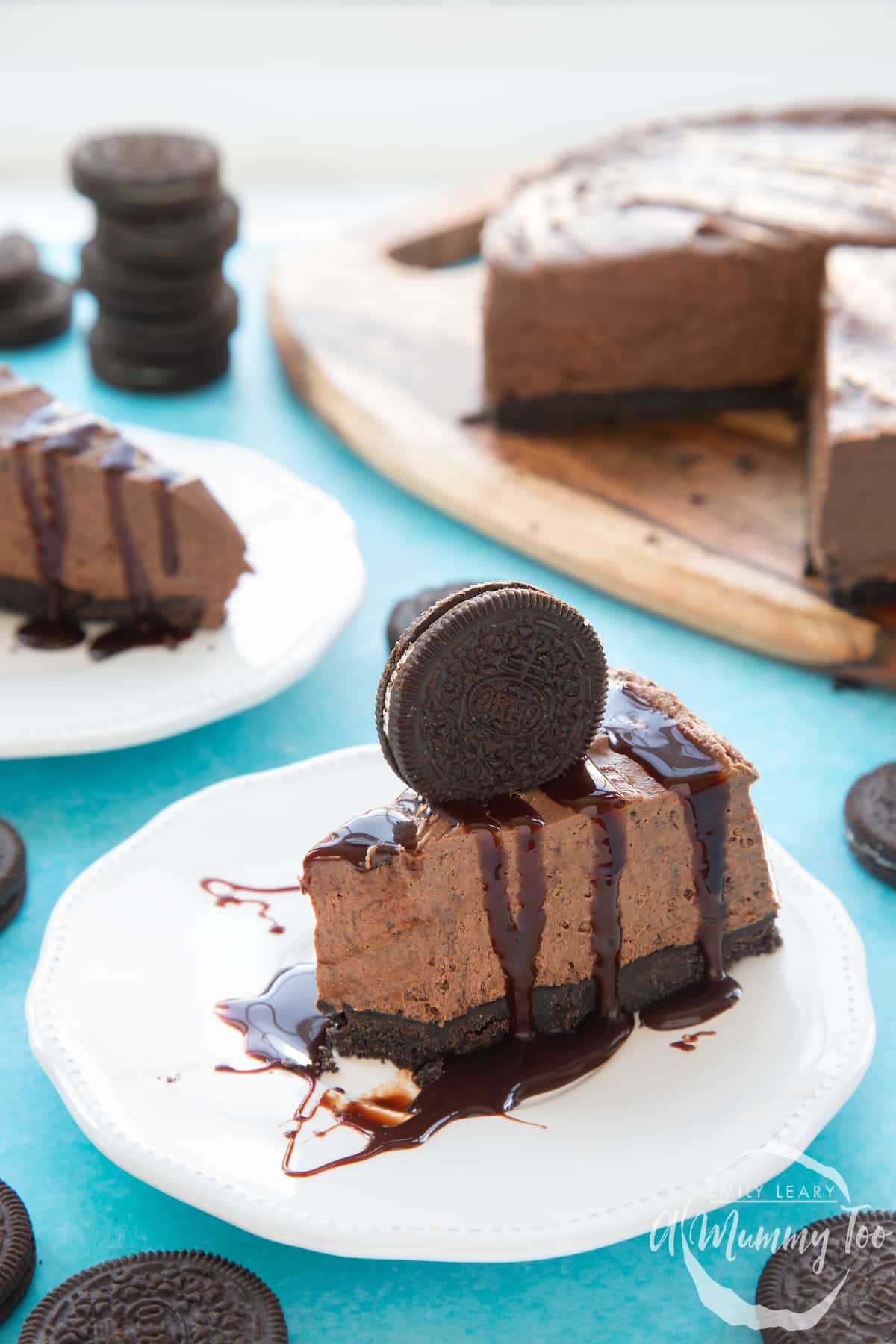 Front view shot of a piece of chocolate oreo cheesecake on a white plate drizzled with melted chocolate sauce topped with a oreo cookie