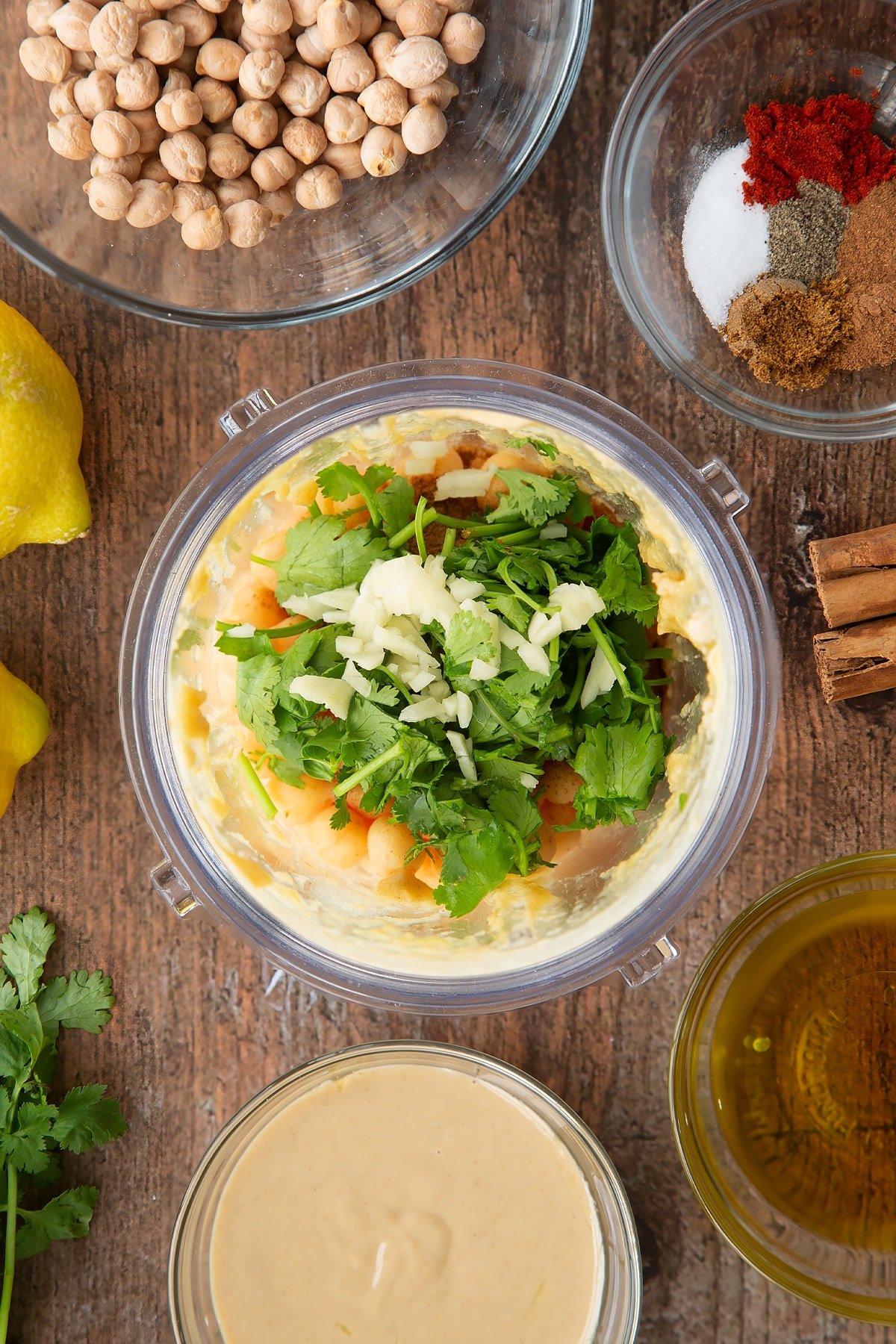 Tahini, lemon juice and oil whipped together in a small blender bowl with cooked chickpeas, spices, fresh coriander and garlic on top. Ingredients to make cinnamon hummus surround the bowl.