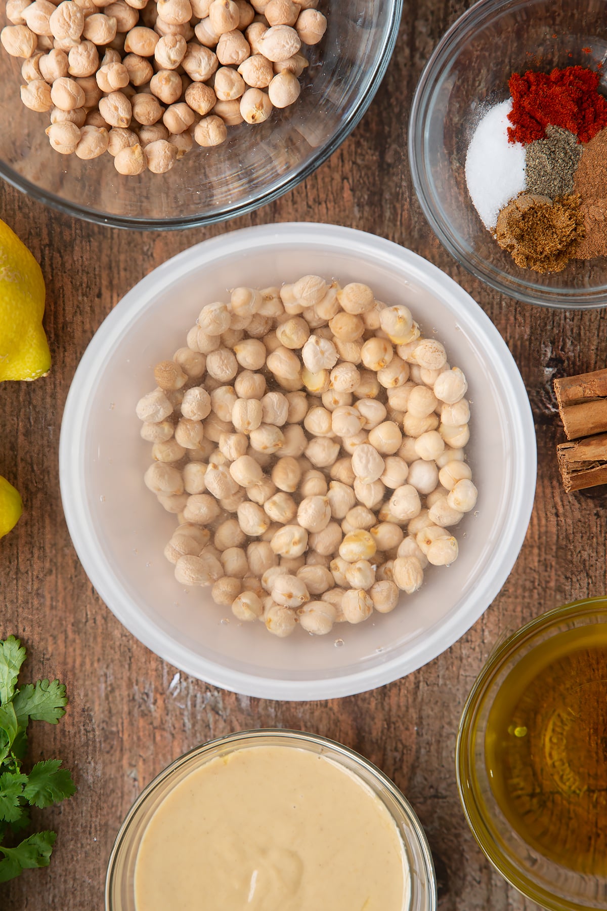Dried chickpeas covered with water in a plastic bowl. Ingredients to make cinnamon hummus surround the bowl.