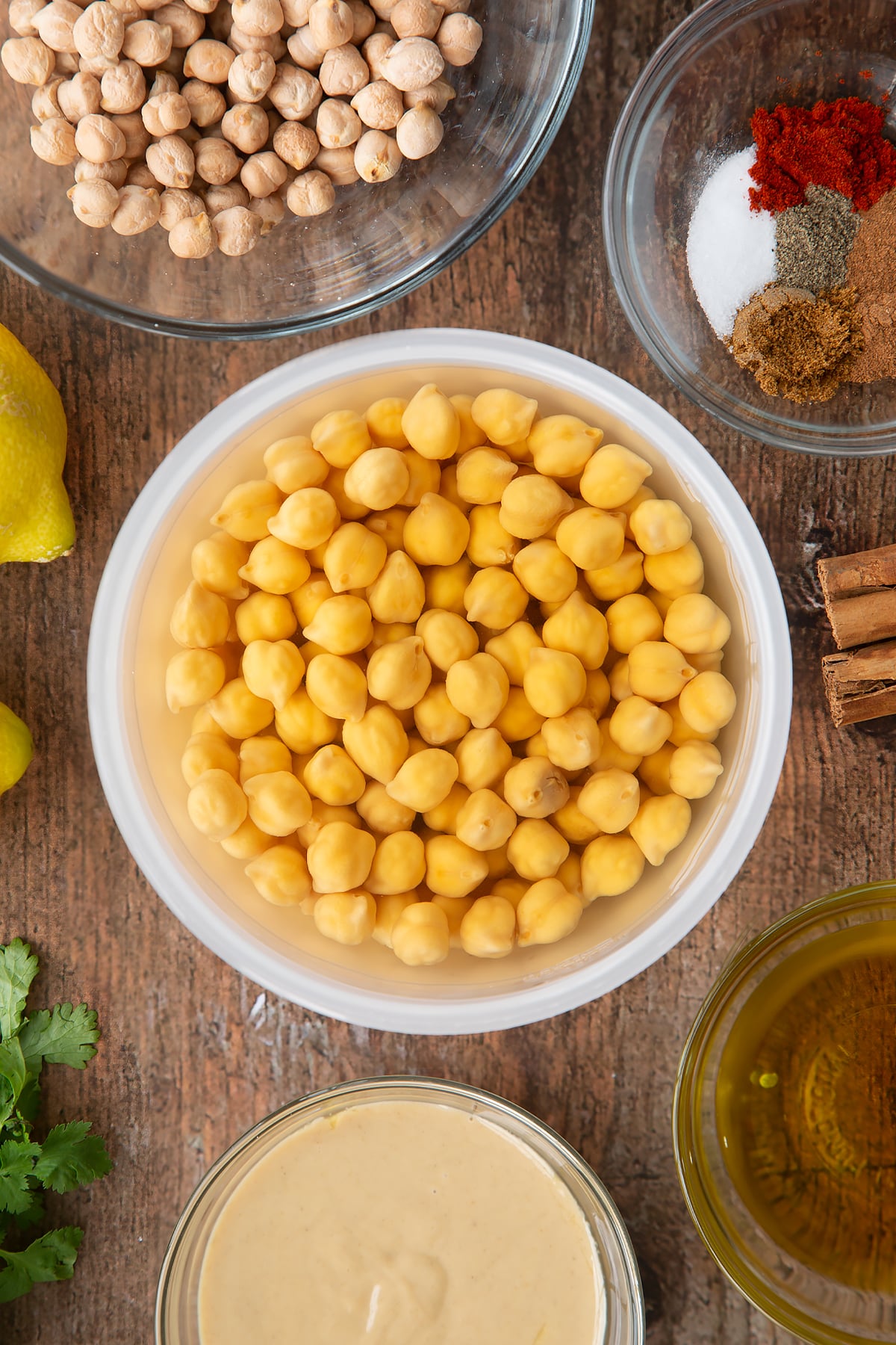 Soaked chickpeas in water in a plastic bowl. Ingredients to make cinnamon hummus surround the bowl.