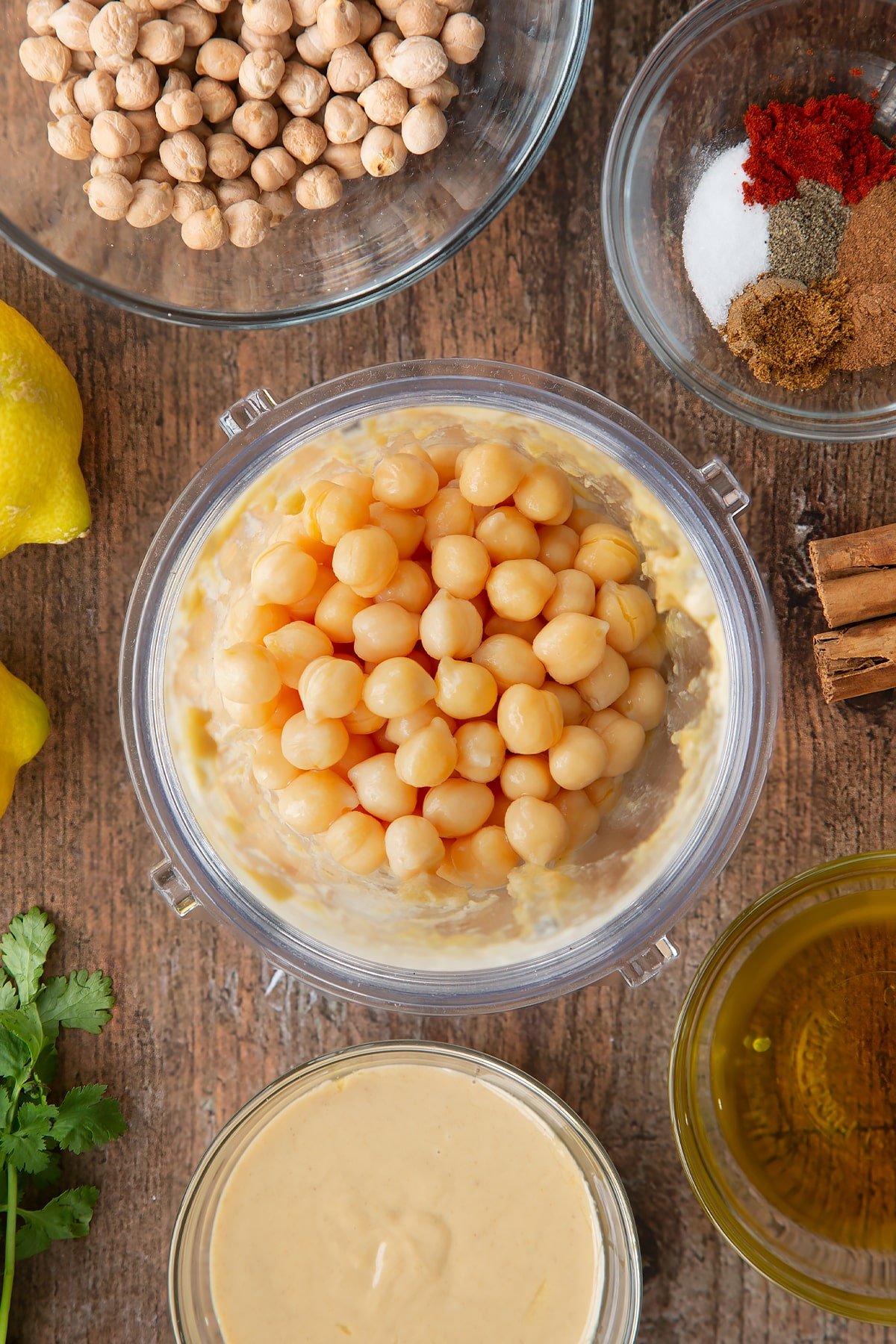 Tahini, lemon juice and oil whipped together in a small blender bowl with cooked chickpeas on top. Ingredients to make cinnamon hummus surround the bowl.