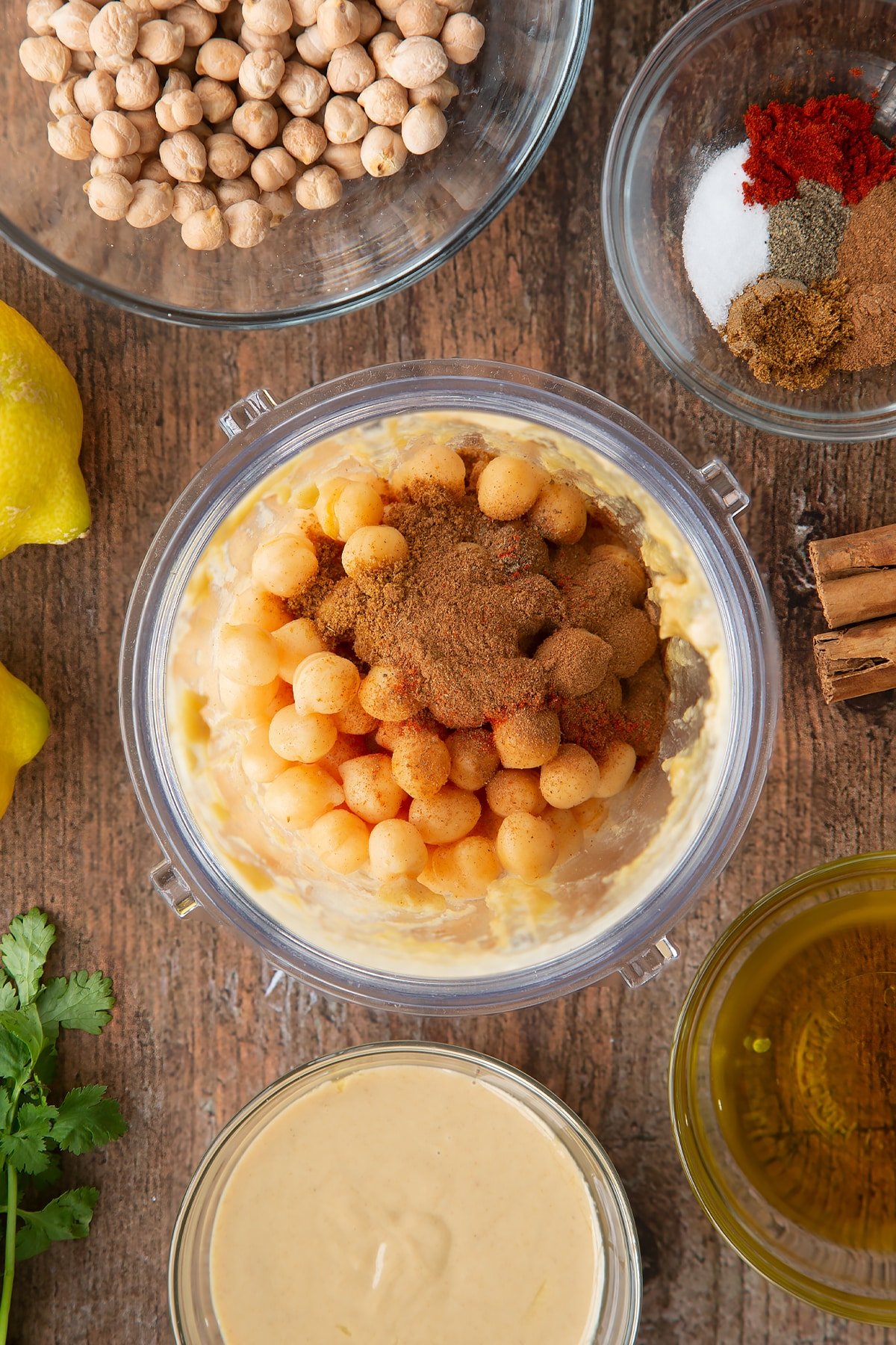 Tahini, lemon juice and oil whipped together in a small blender bowl with cooked chickpeas and spices on top. Ingredients to make cinnamon hummus surround the bowl.