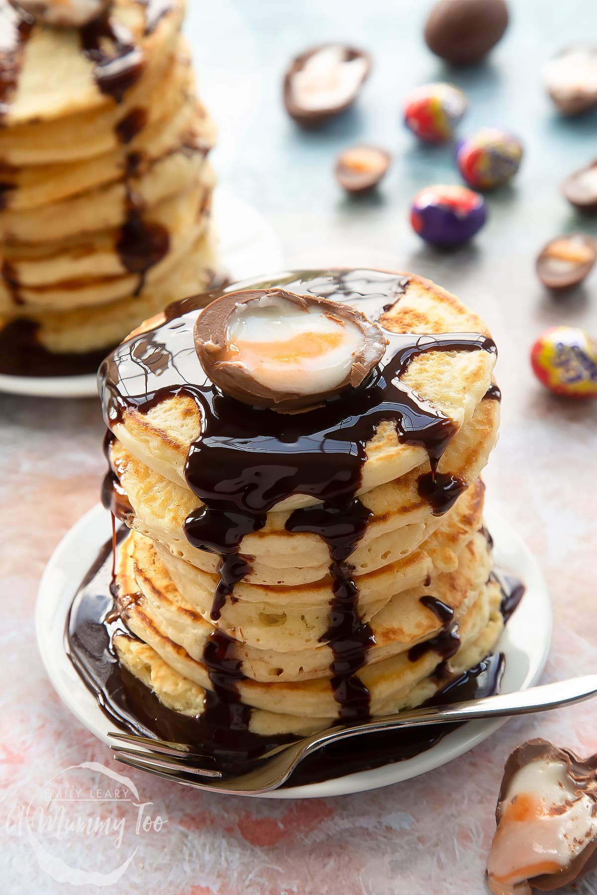 A tall stack of Creme Egg pancakes on a small white plate with a fork. The stack is topped with chocolate sauce and a Creme Egg half. 