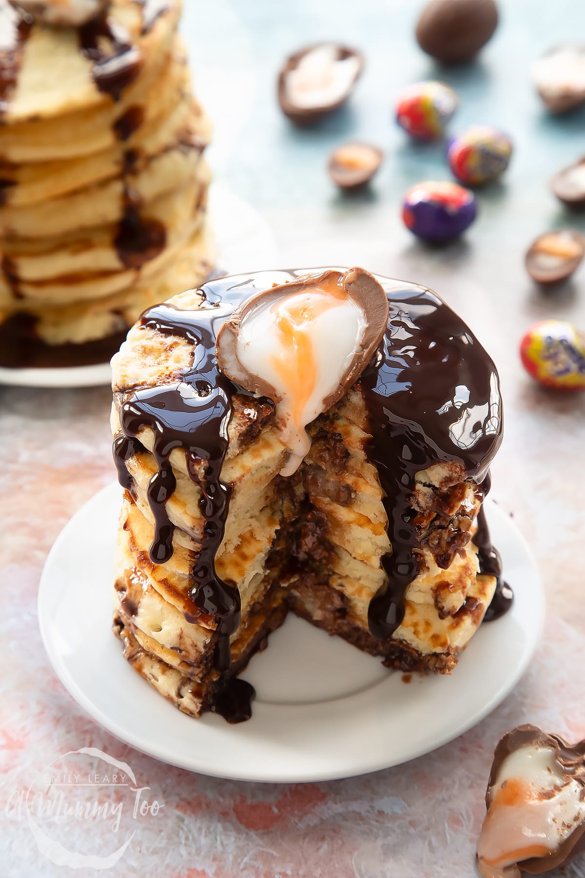 A tall stack of Creme Egg pancakes on a small white plate. The stack is topped with chocolate sauce and a Creme Egg half. It has a wedge cut out of it.