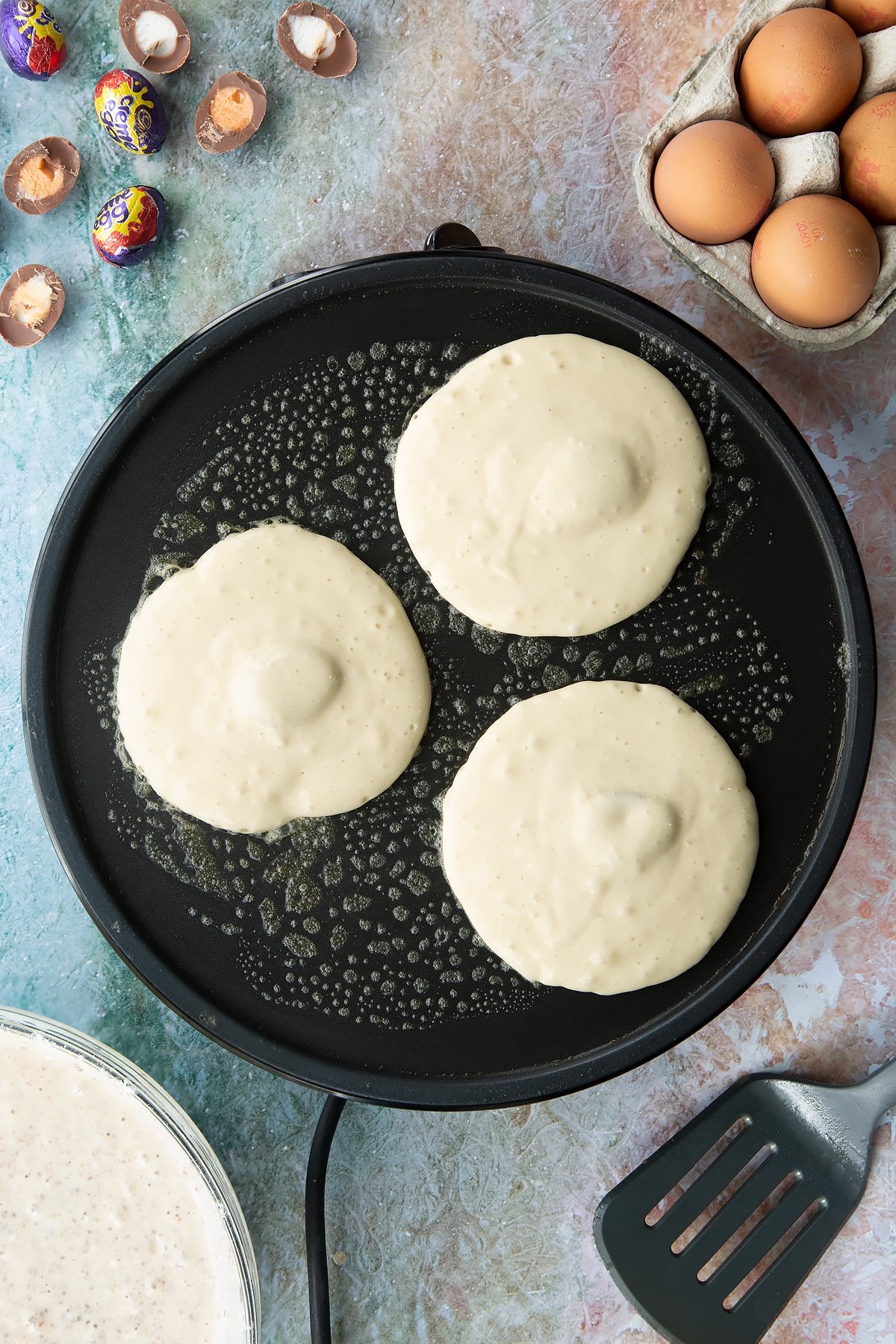 A hot pan with three scoops of pancake batter stuffed with Creme Egg mini halves. Ingredients to make Creme Egg pancakes surround the pan.