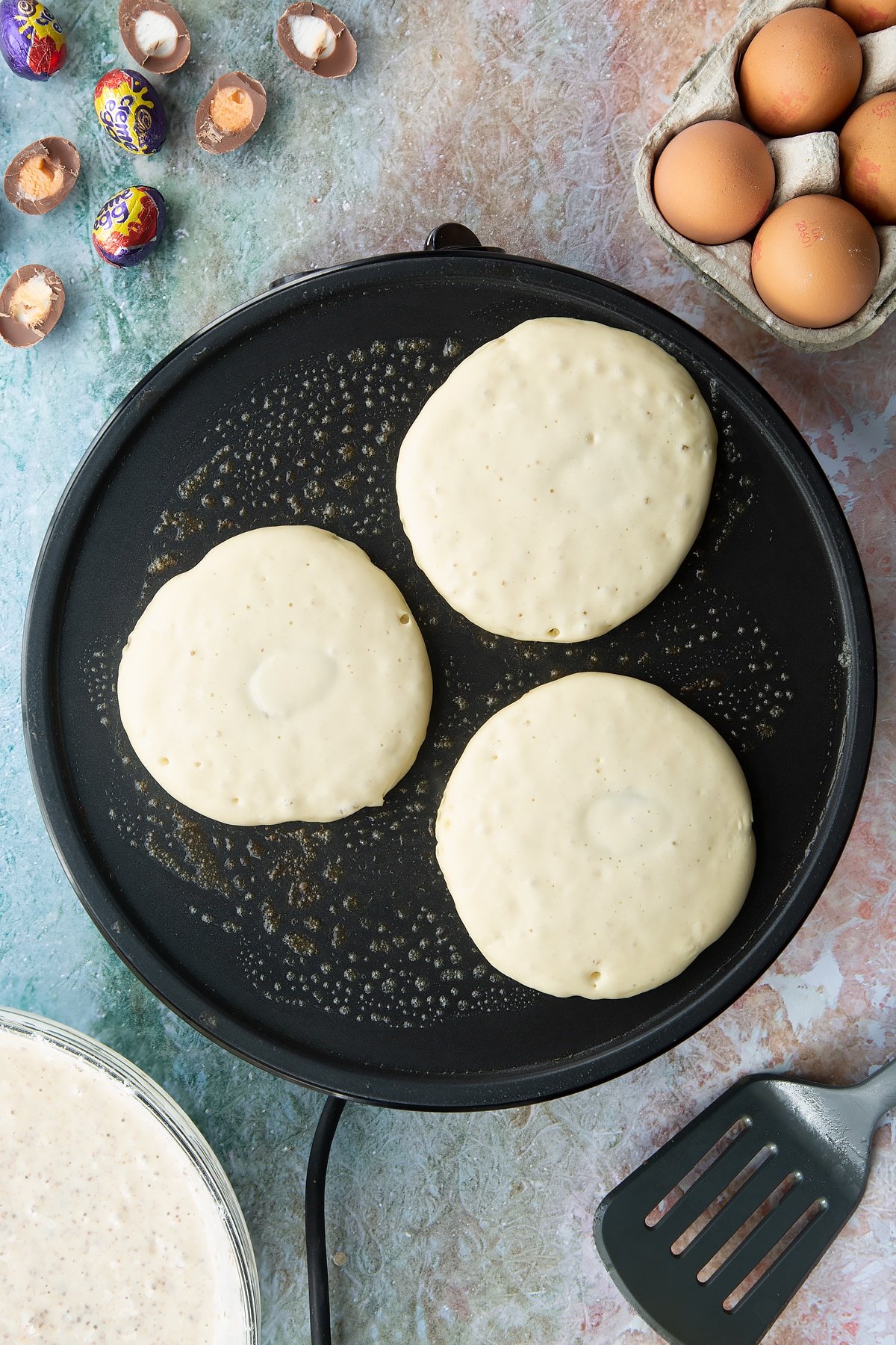 A hot pan with three scoops of half cooked pancake batter stuffed with Creme Egg mini halves. Ingredients to make Creme Egg pancakes surround the pan.