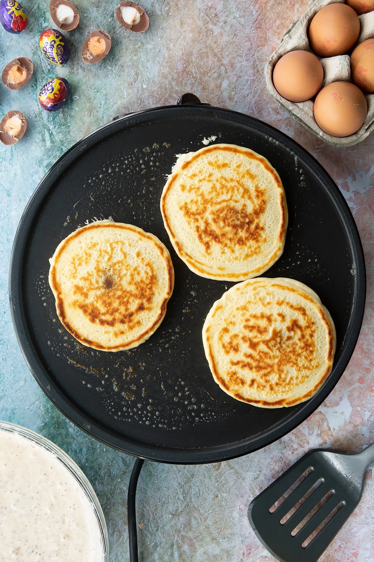 A hot pan with Creme Egg pancakes cooking. Ingredients to make Creme Egg pancakes surround the pan.