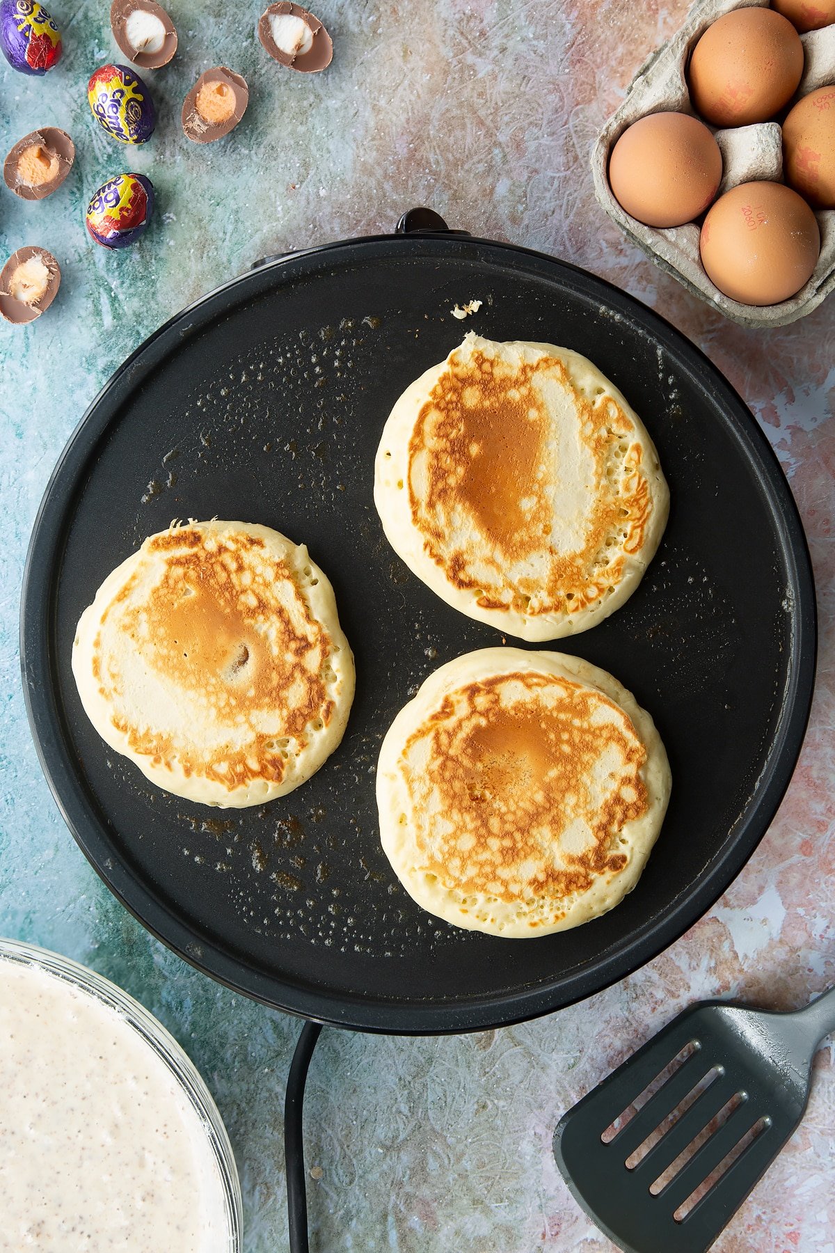 A hot pan with Creme Egg pancakes fully cooked. Ingredients to make Creme Egg pancakes surround the pan.