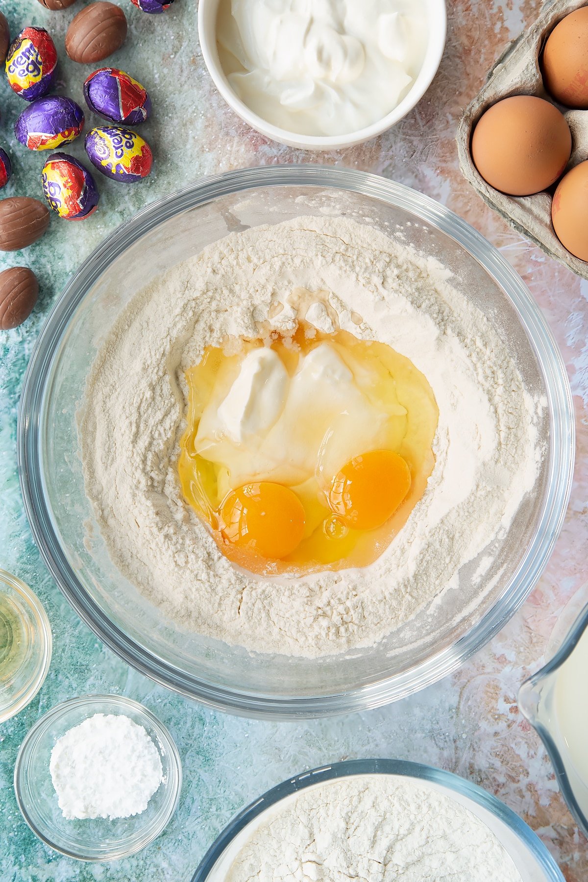 Self-raising flour and baking powder in a bowl with a well in the centre filled with crème fraîche, eggs and vanilla. Ingredients to make Creme Egg pancakes surround the bowl.