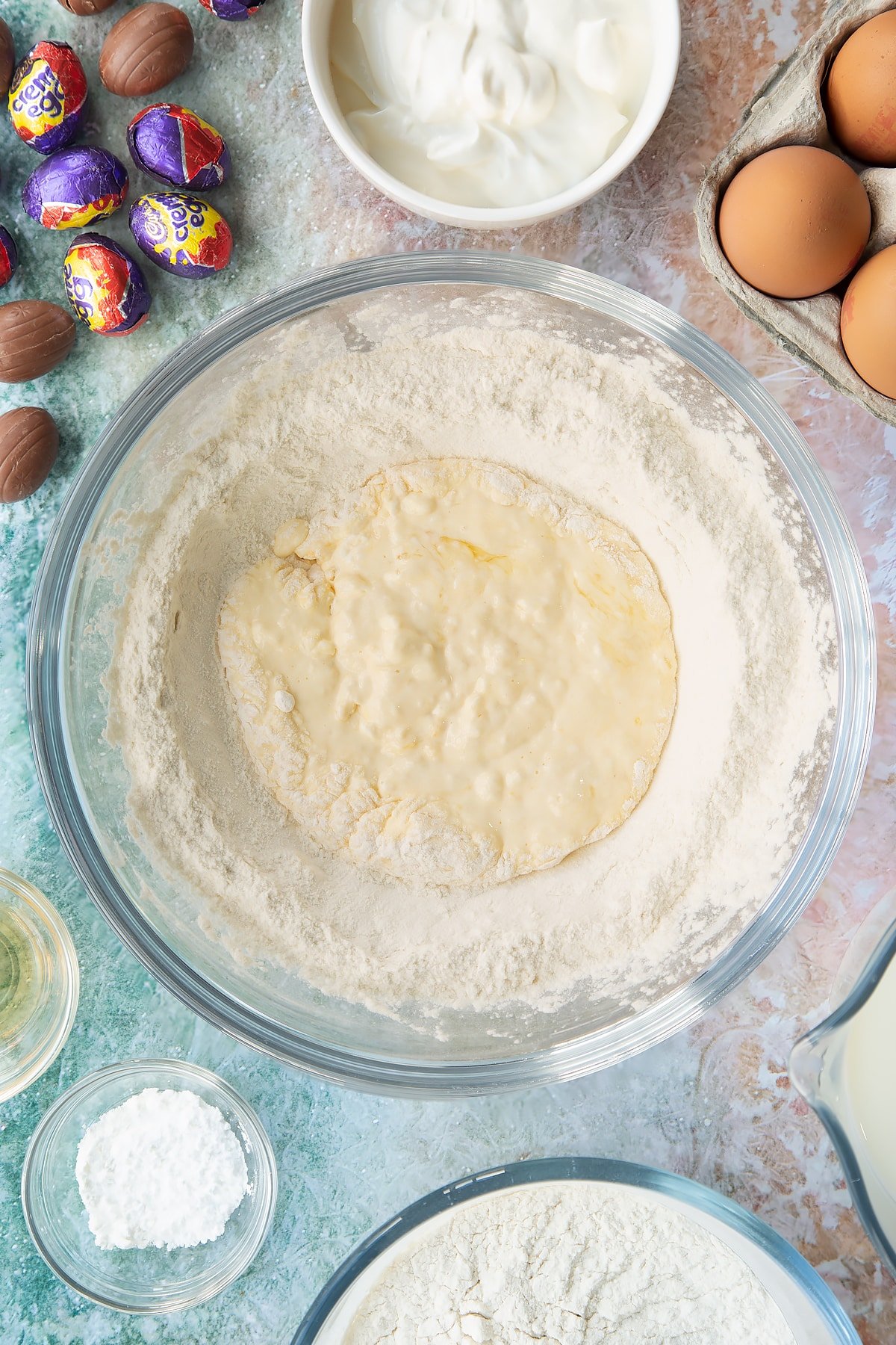 Self-raising flour and baking powder in a bowl with a well in the centre filled with crème fraîche, eggs and vanilla, with some of the flour incorporated. Ingredients to make Creme Egg pancakes surround the bowl.