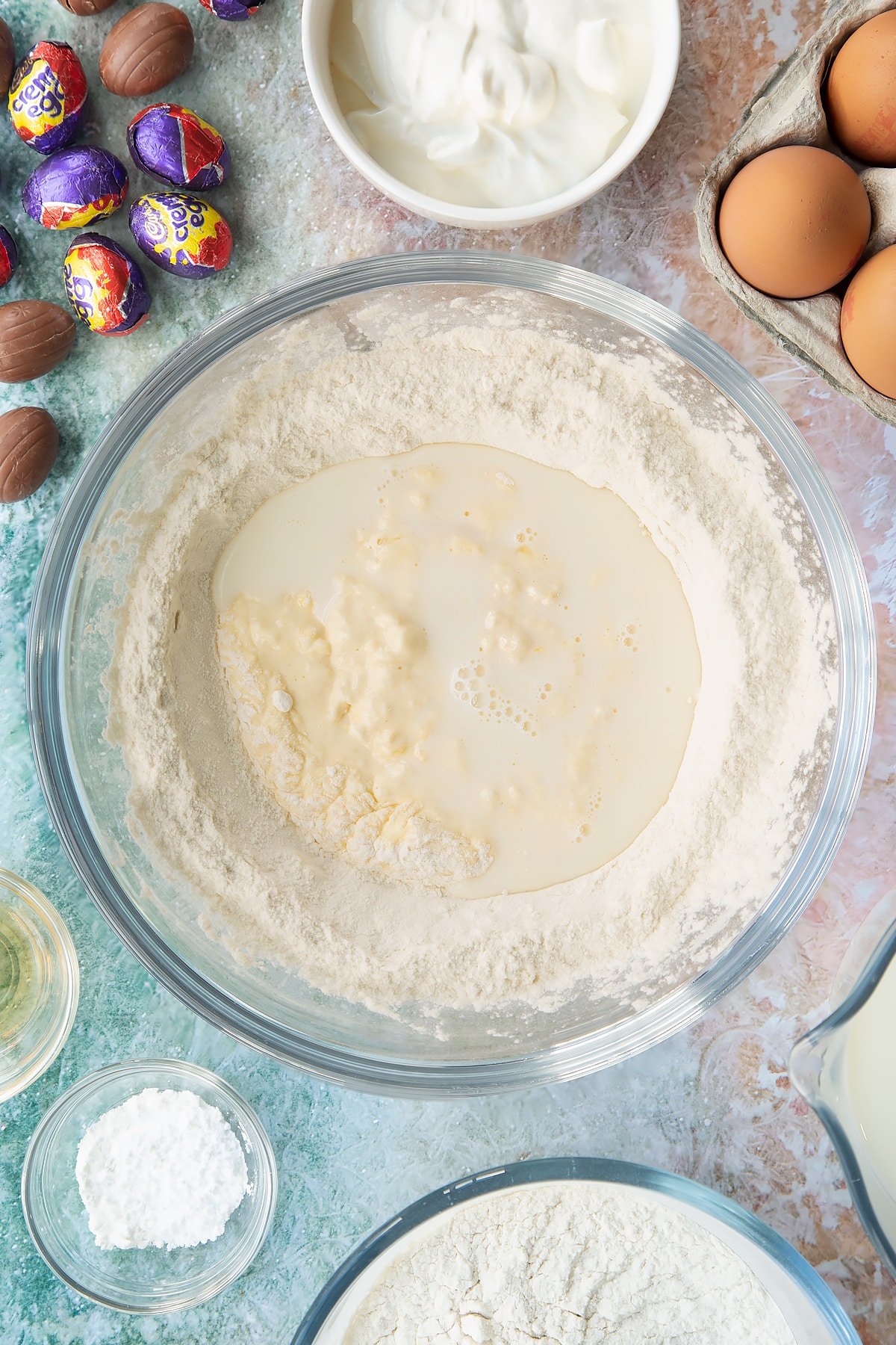 Self-raising flour and baking powder in a bowl with a well in the centre filled with crème fraîche, eggs, vanilla and milk, with some of the flour incorporated. Ingredients to make Creme Egg pancakes surround the bowl.
