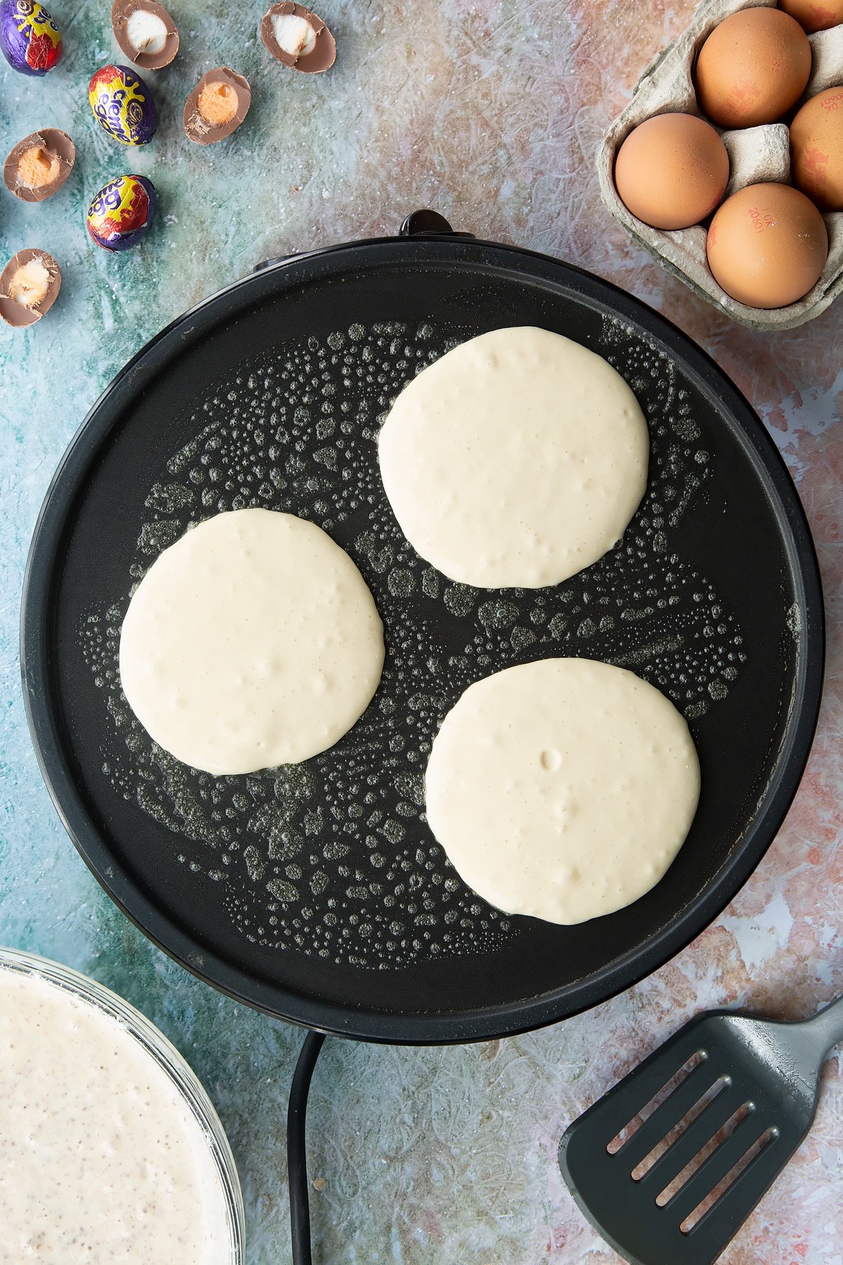 A hot pan with three scoops of pancake batter. Ingredients to make Creme Egg pancakes surround the pan.
