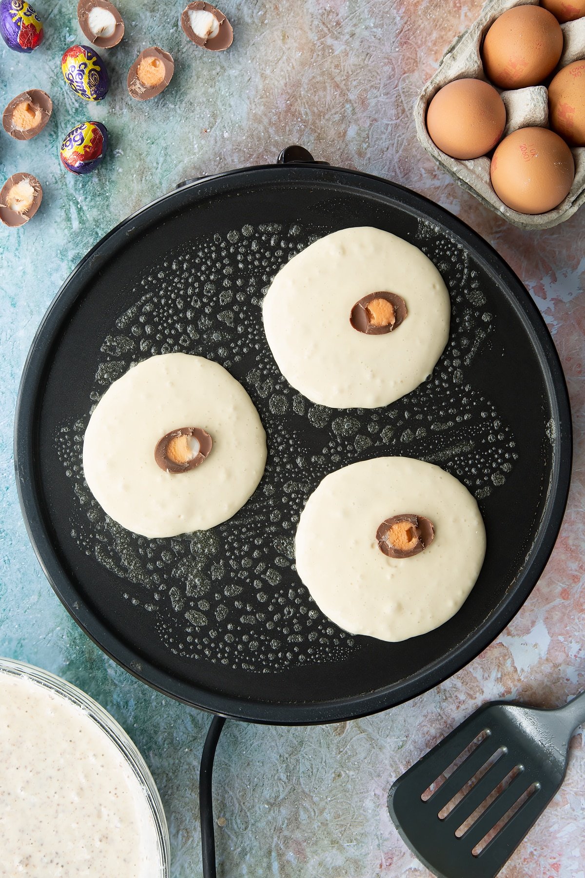 A hot pan with three scoops of pancake batter topped with Creme Egg mini halves. Ingredients to make Creme Egg pancakes surround the pan.