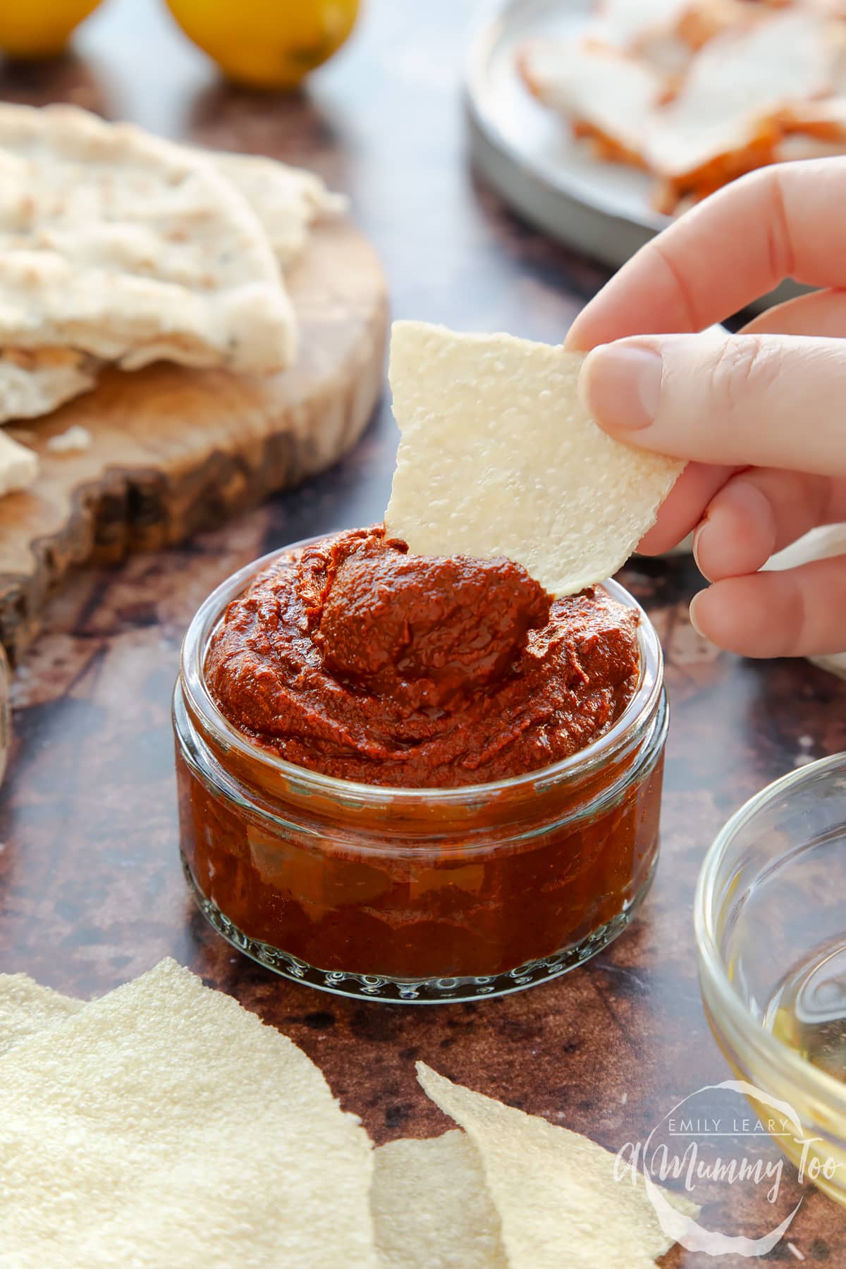 Front view of a hand holding a piece of bread dipping into garlic chutney in a small glass jar with the A Mummy Too logo in the corner