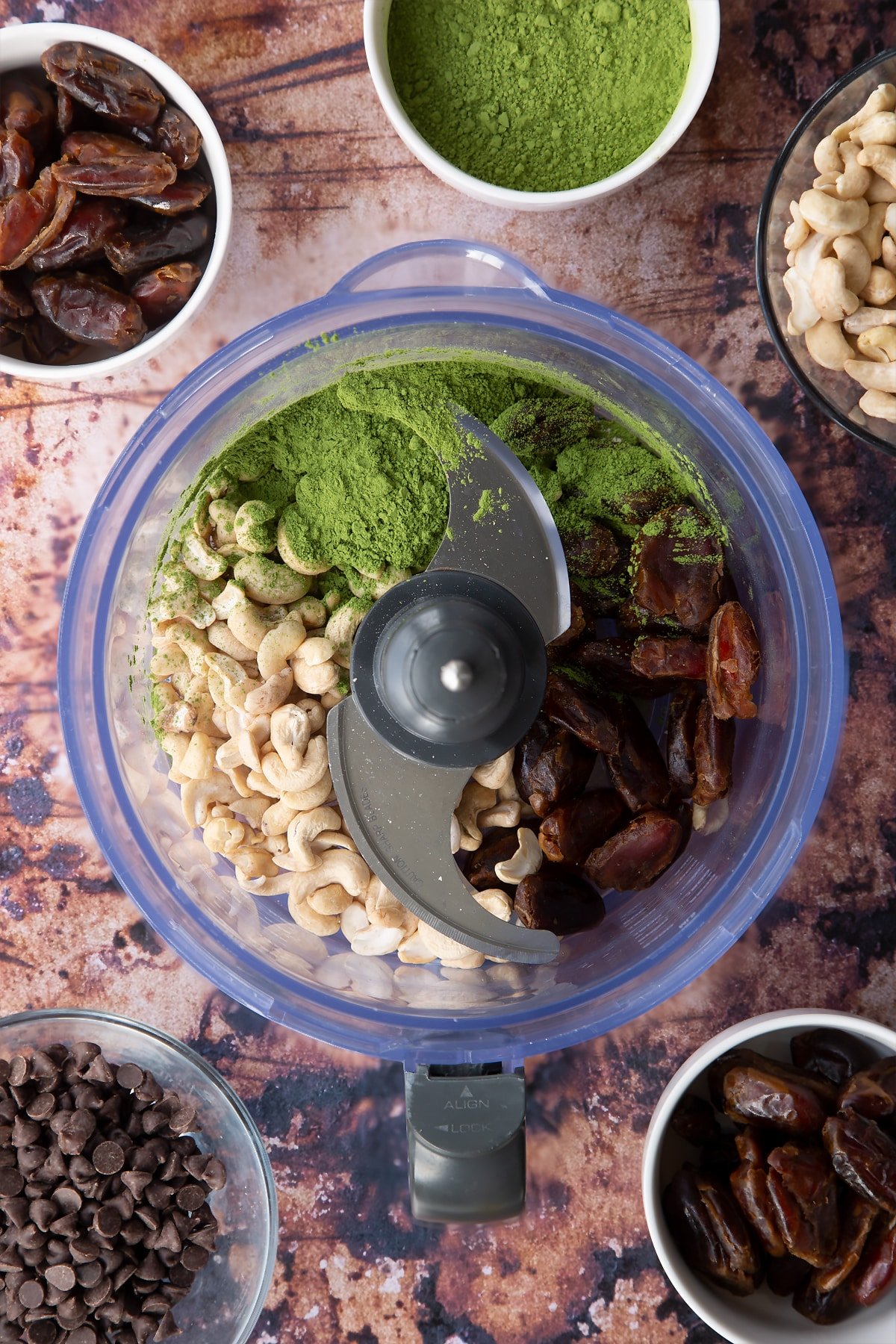 Dates, cashews and matcha green tea powder in a blender bowl. Ingredients to make matcha protein balls surround the bowl.