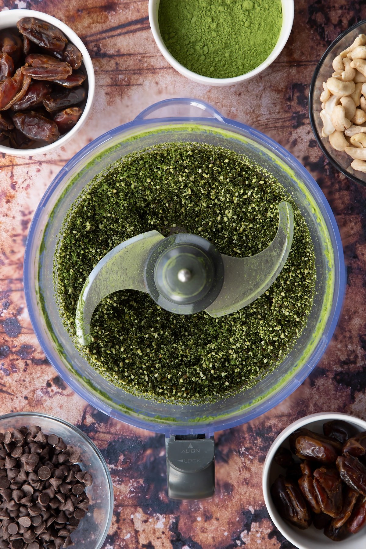 Dates, cashews and matcha green tea powder blended to a coarse crumb in a blender bowl. Ingredients to make matcha protein balls surround the bowl.