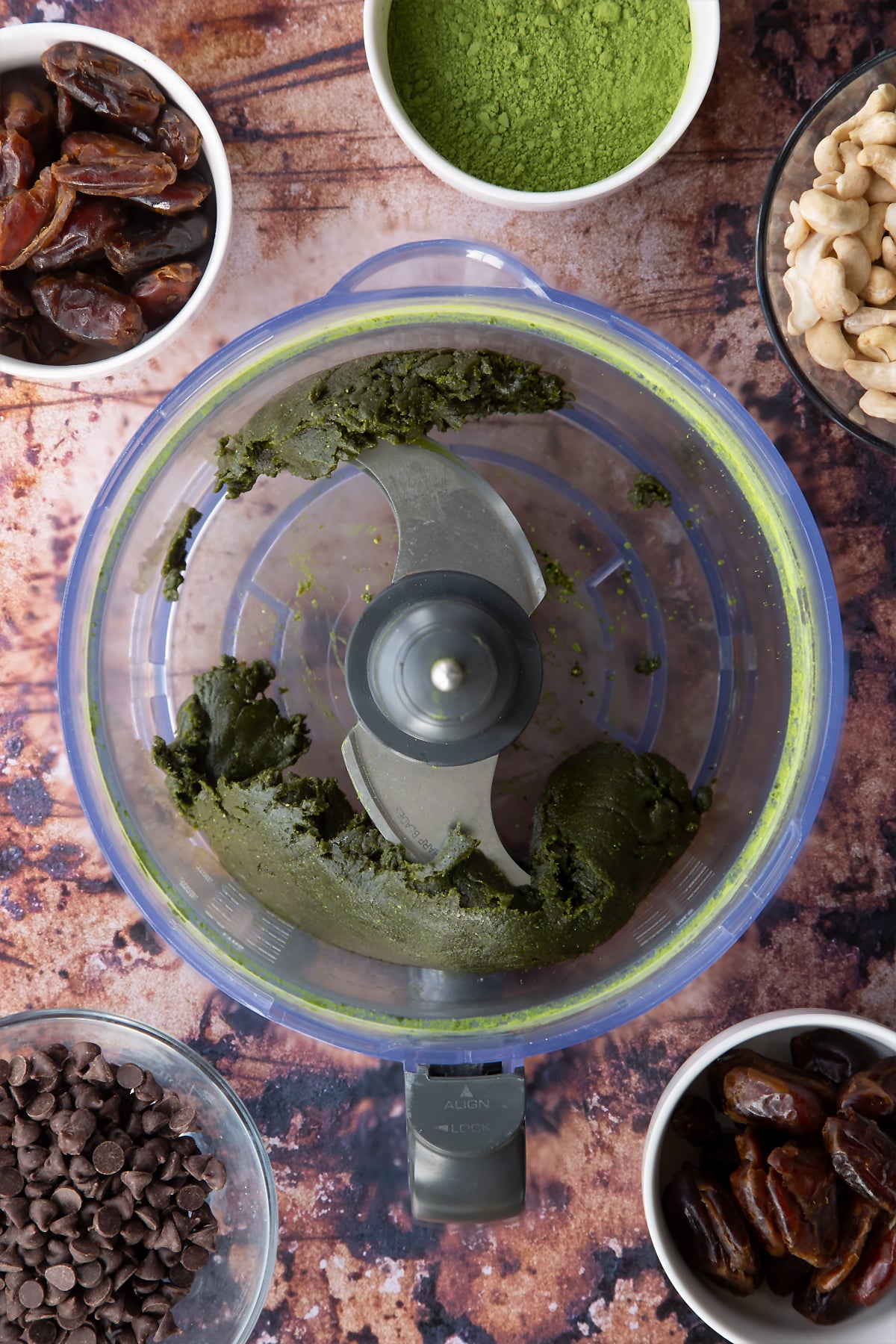 Dates, cashews and matcha green tea powder blended to a paste in a blender bowl. Ingredients to make matcha protein balls surround the bowl.