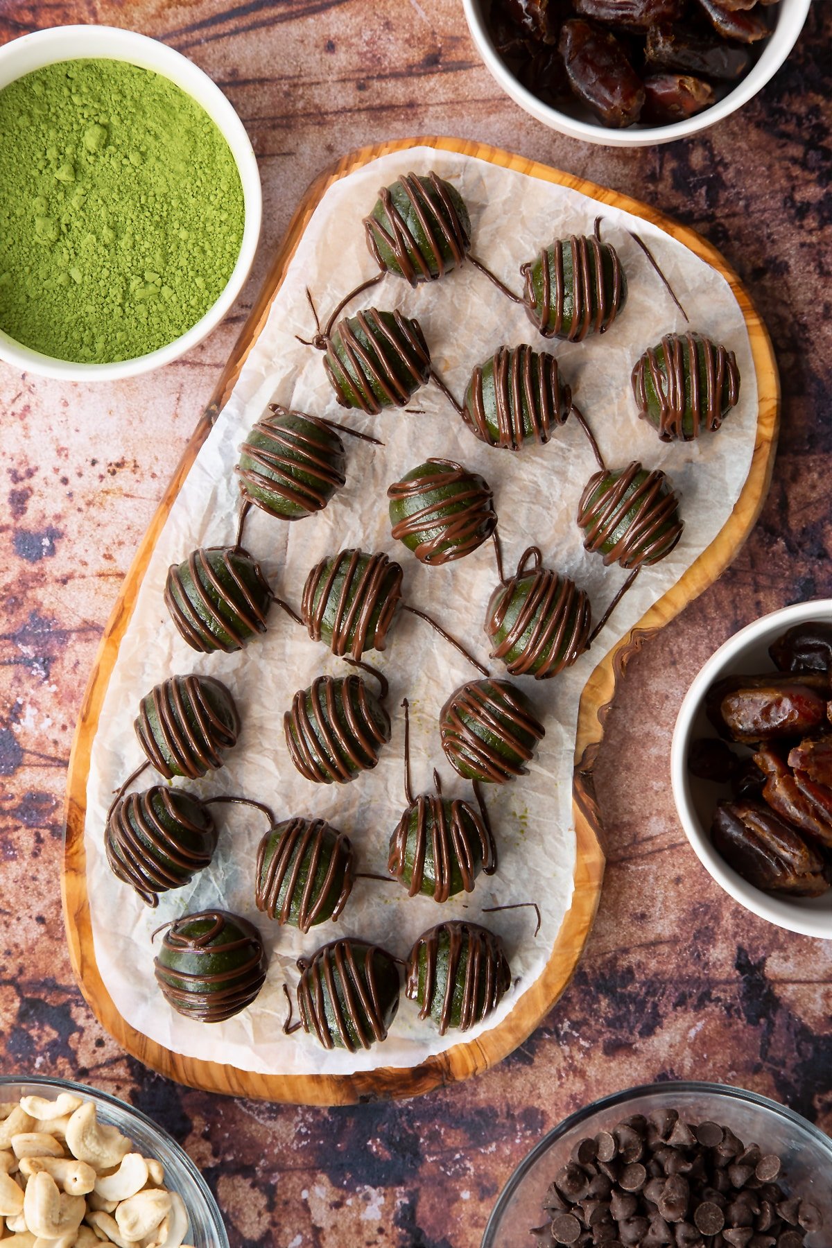 Matcha protein balls shown from above on a wooden board lined with baking paper. The balls are dark green and have been drizzled with dark chocolate.