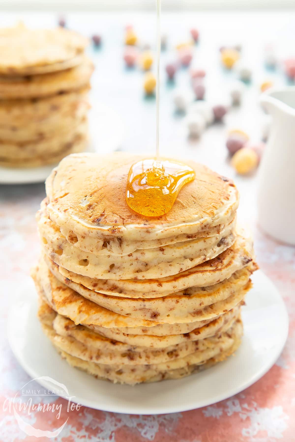 Thick, fluffy Mini Egg pancakes on a white plate in a tall stack. They are being drizzled with syrup.