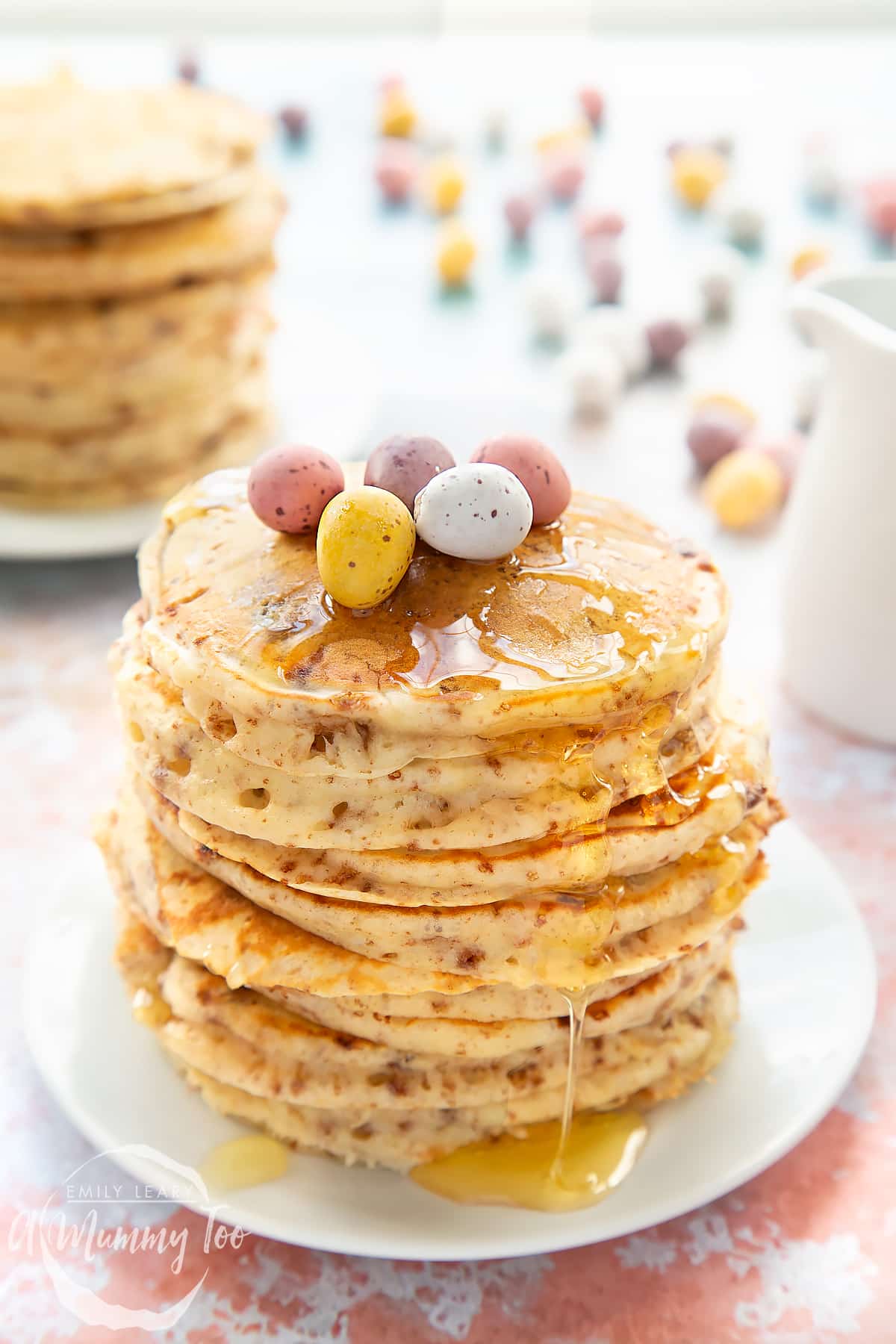 Thick, fluffy Mini Egg pancakes on a white plate in a tall stack. They are drizzled with syrup and topped with Mini Eggs.