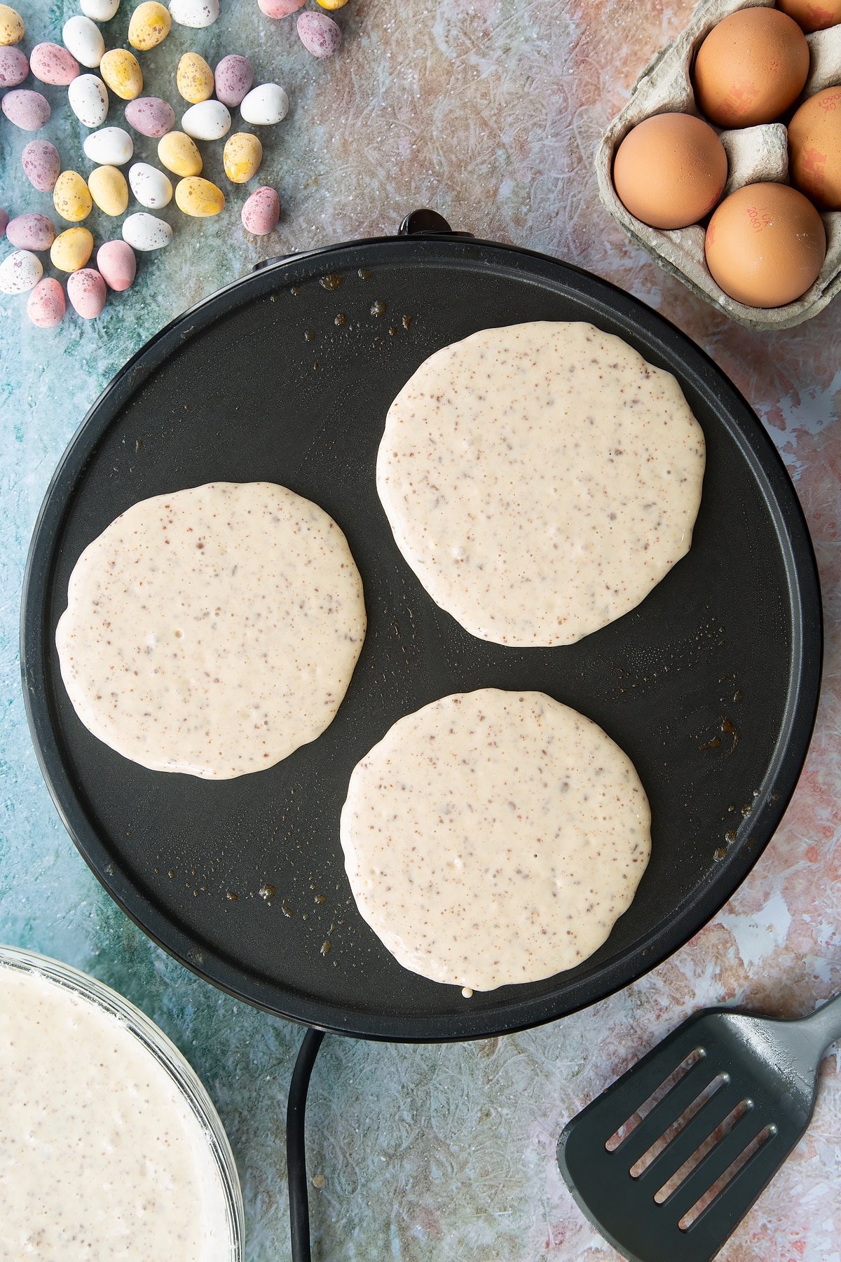 Spoonfuls of Mini Egg pancake batter on a hot pan. Ingredients to make Mini Egg pancakes surround the pan.