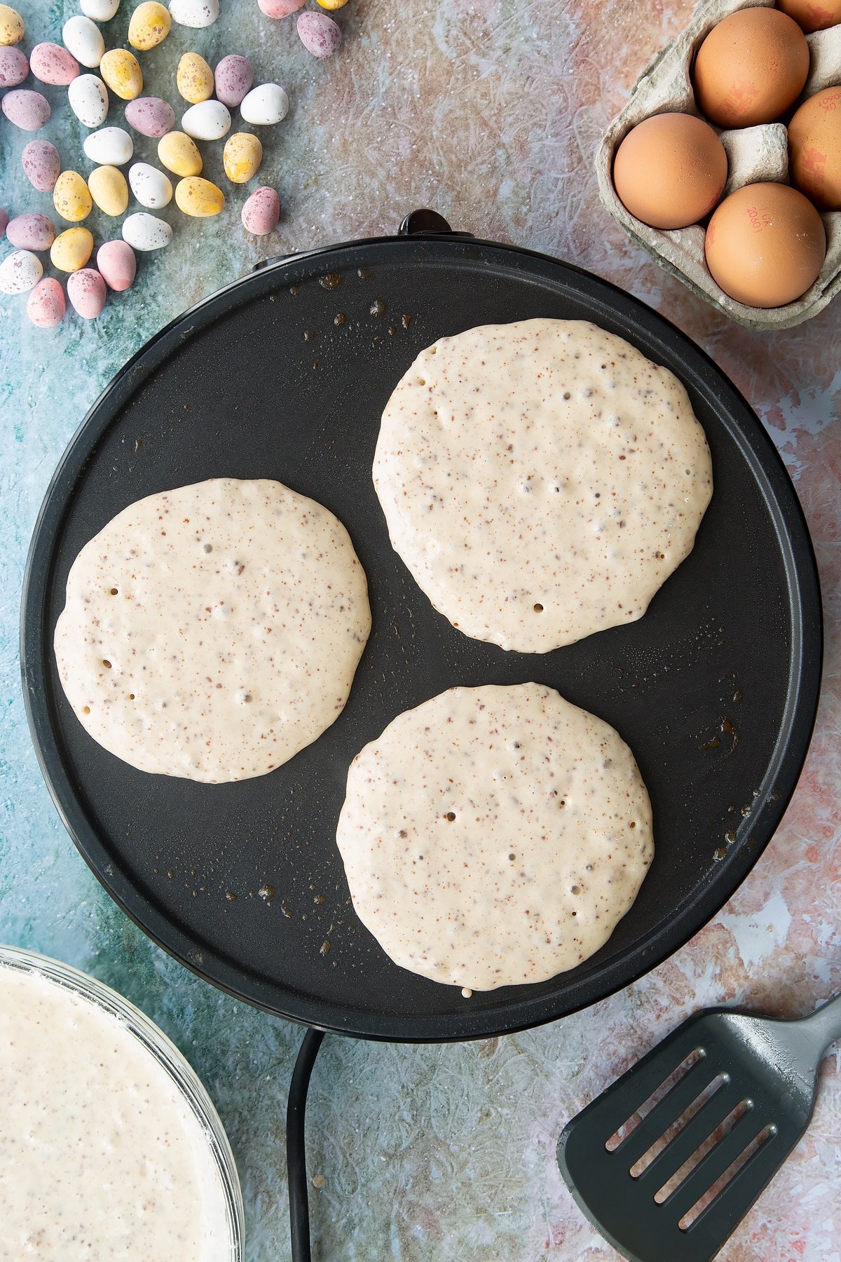Spoonfuls of bubbling Mini Egg pancake batter on a hot pan. Ingredients to make Mini Egg pancakes surround the pan.