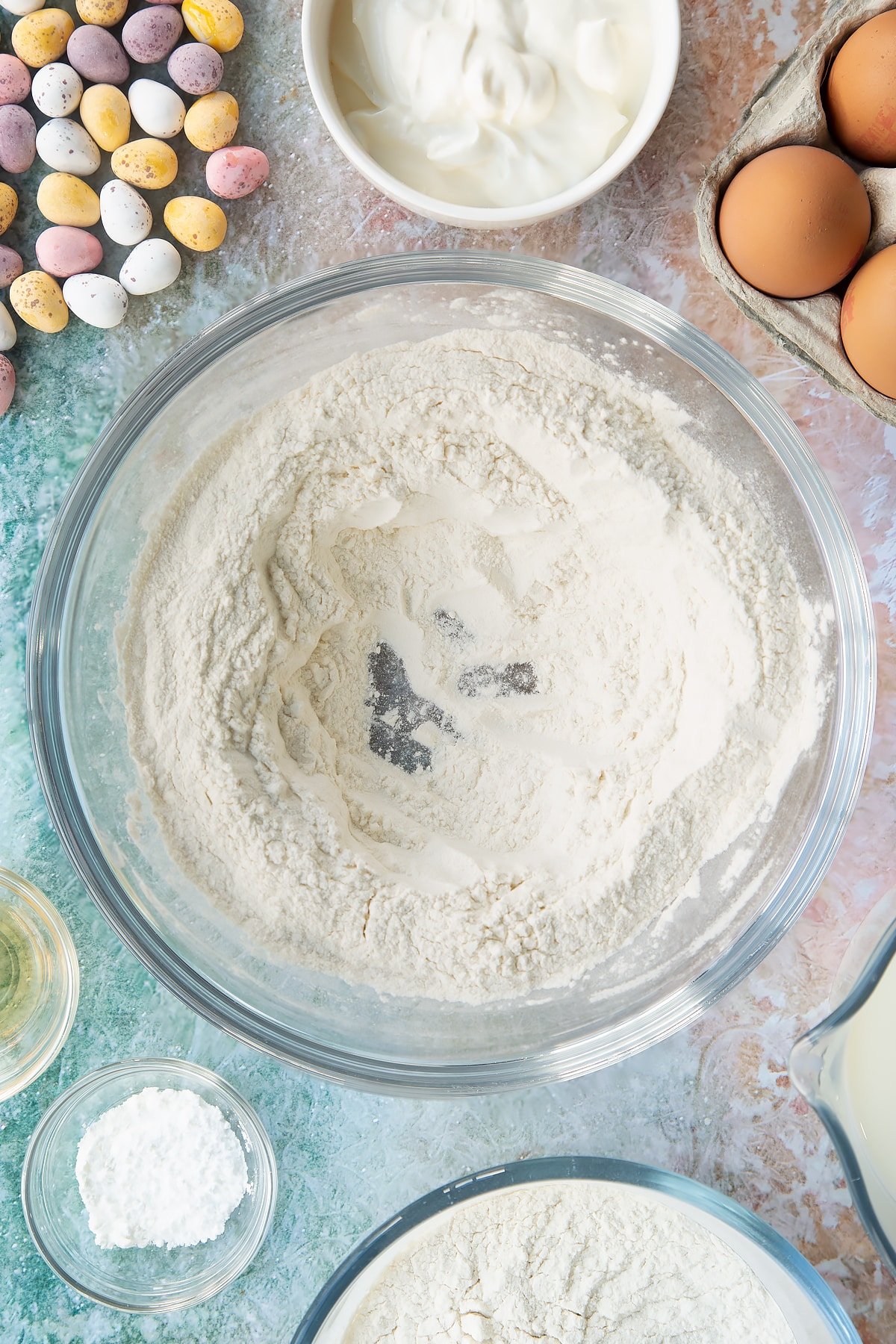 Flour and baking powder in a bowl with a well in the centre. Ingredients to make Mini Egg pancakes surround the bowl.