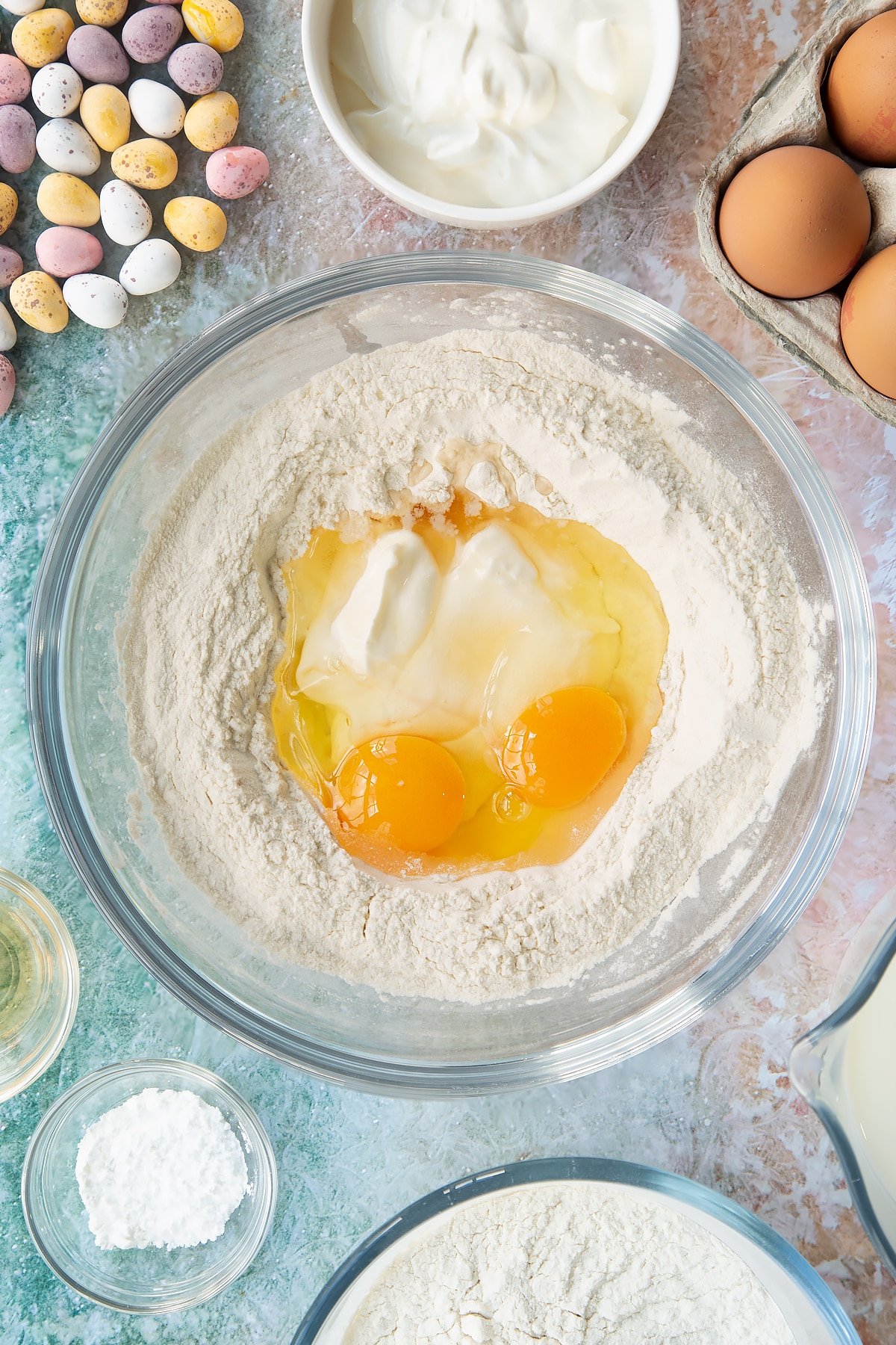 Flour and baking powder in a bowl with a well in the centre filled with creme fraiche and eggs. Ingredients to make Mini Egg pancakes surround the bowl.