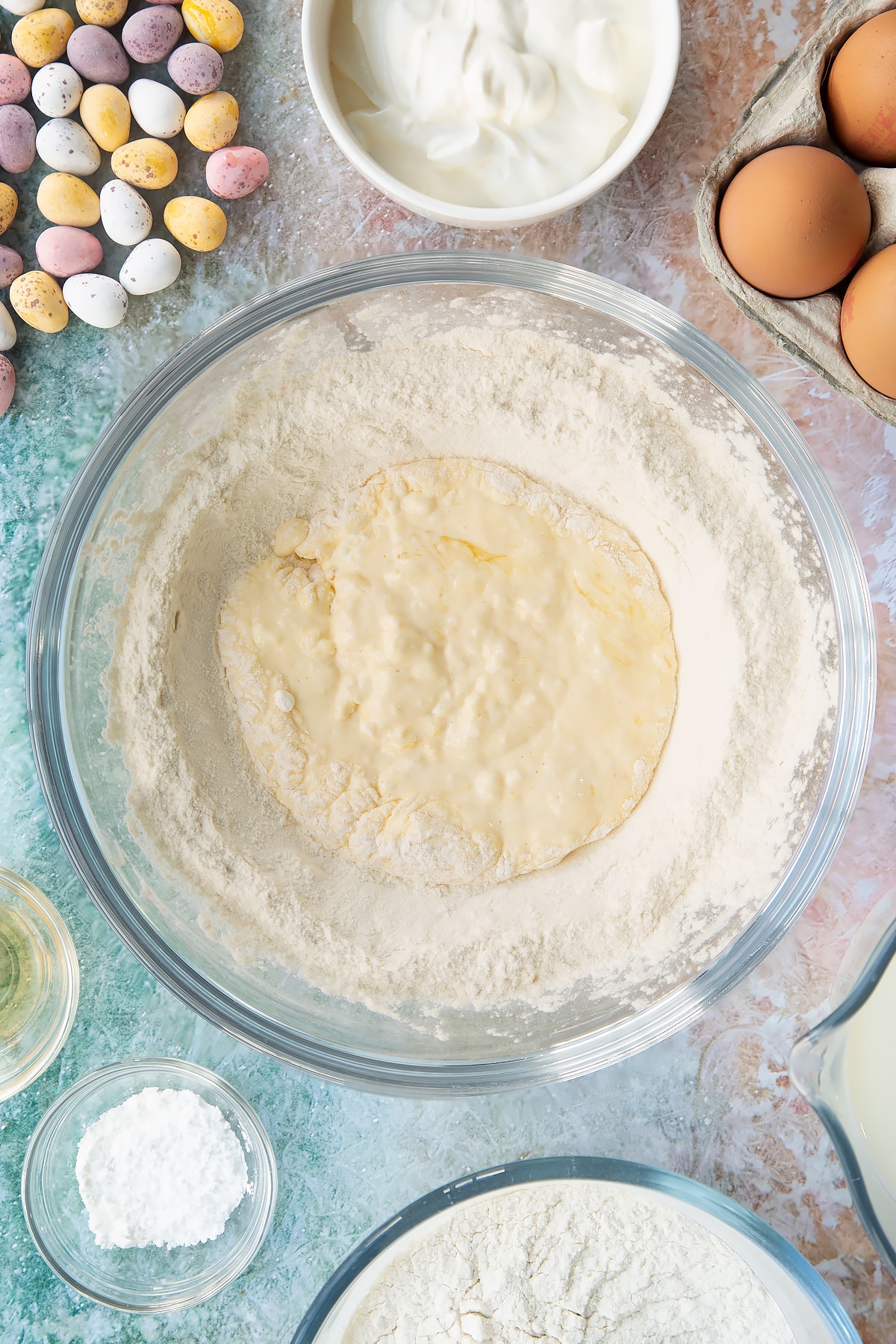 Flour and baking powder in a bowl with a well in the centre filled with whisked creme fraiche and eggs. Ingredients to make Mini Egg pancakes surround the bowl.