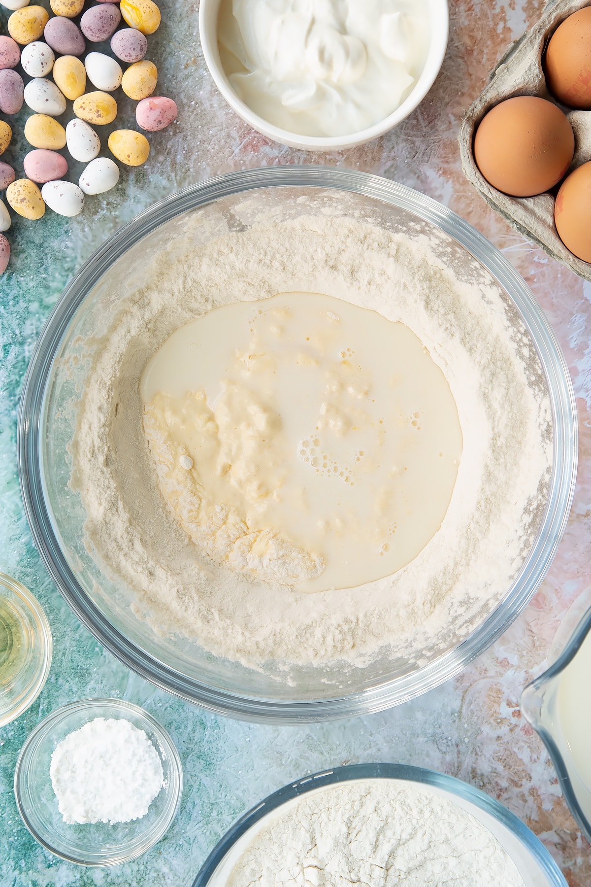 Flour and baking powder in a bowl with a well in the centre filled with whisked creme fraiche, milk and eggs. Ingredients to make Mini Egg pancakes surround the bowl.