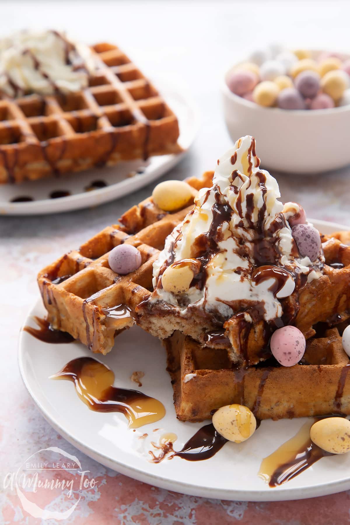 Mini egg waffles on a white plate. One has been cut in to. They are topped with squirty cream, chocolate sauce, toffee sauce and Mini Eggs.