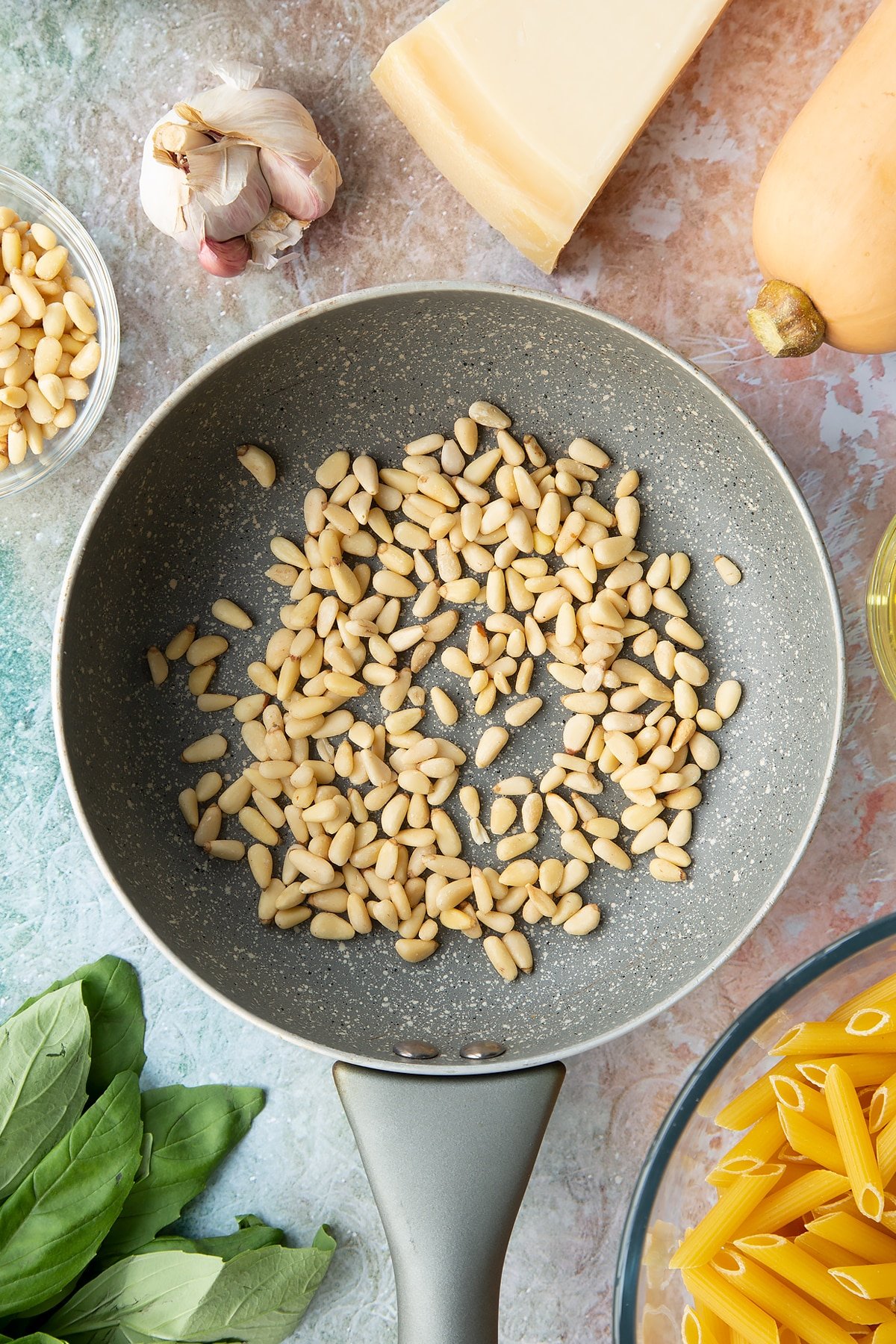 Pine nuts in a small frying pan. Ingredients to make penne with butternut squash and goat cheese surround the pan.