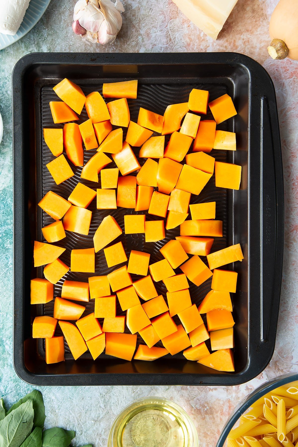 Chunks of butternut squash on a black tray. Ingredients to make penne with butternut squash and goat cheese surround the tray.