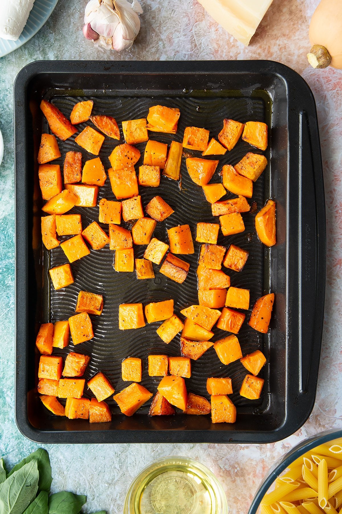 Roasted chunks of butternut squash on a black tray. Ingredients to make penne with butternut squash and goat cheese surround the tray.