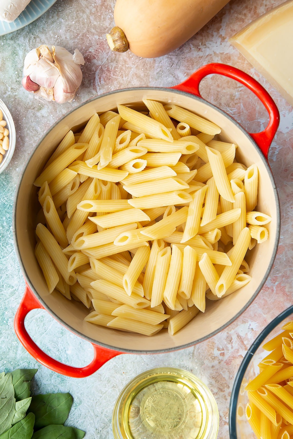 Cooked and drained penne pasta in a pan. Ingredients to make penne with butternut squash and goat cheese surround the pan.