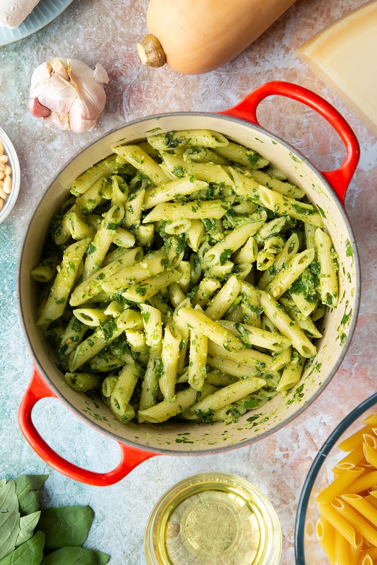 Penne pasta tossed with fresh pesto in a pan. Ingredients to make penne with butternut squash and goat cheese surround the pan.