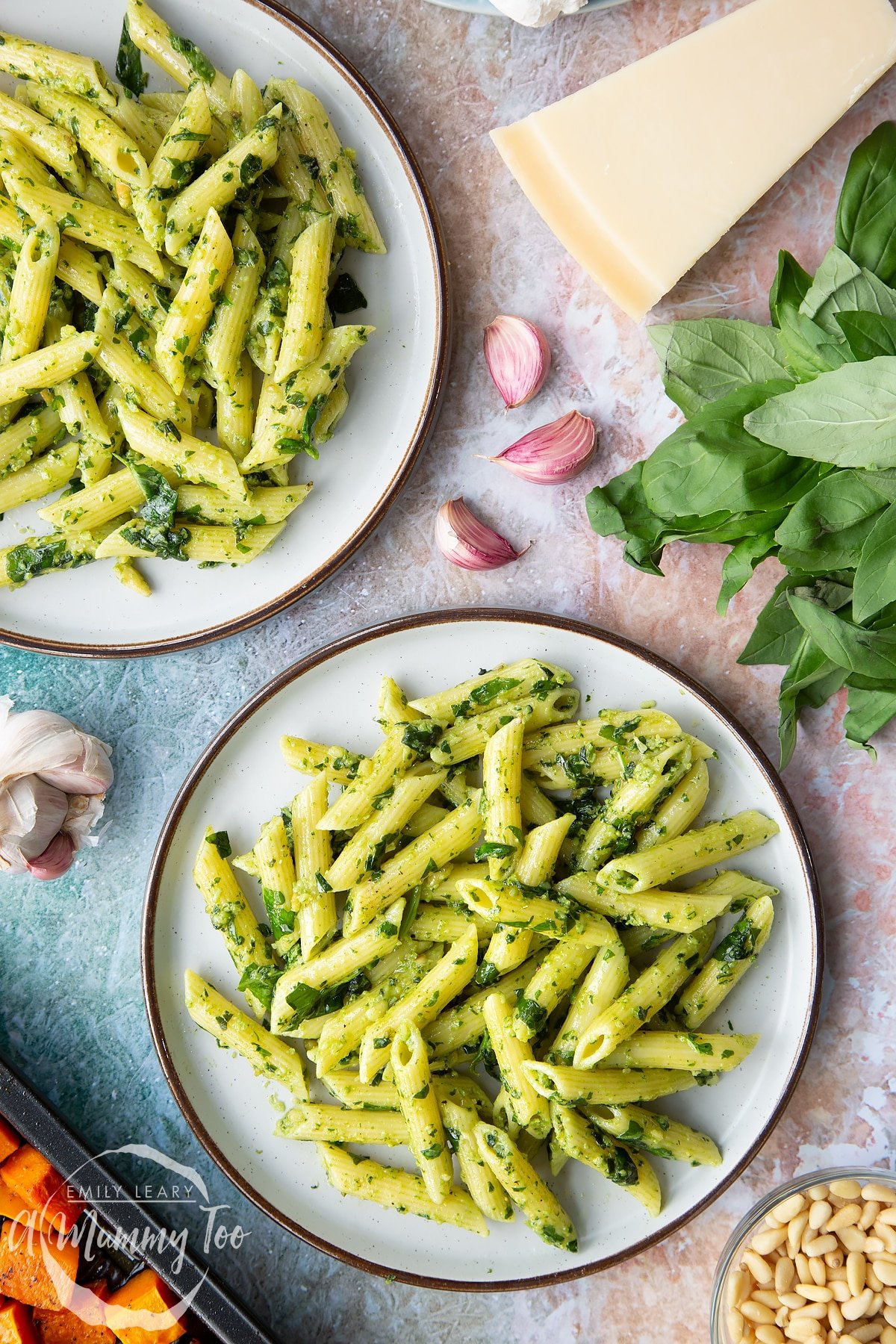 Penne pasta tossed with fresh pesto served to plates. Ingredients to make penne with butternut squash and goat cheese surround the plates.