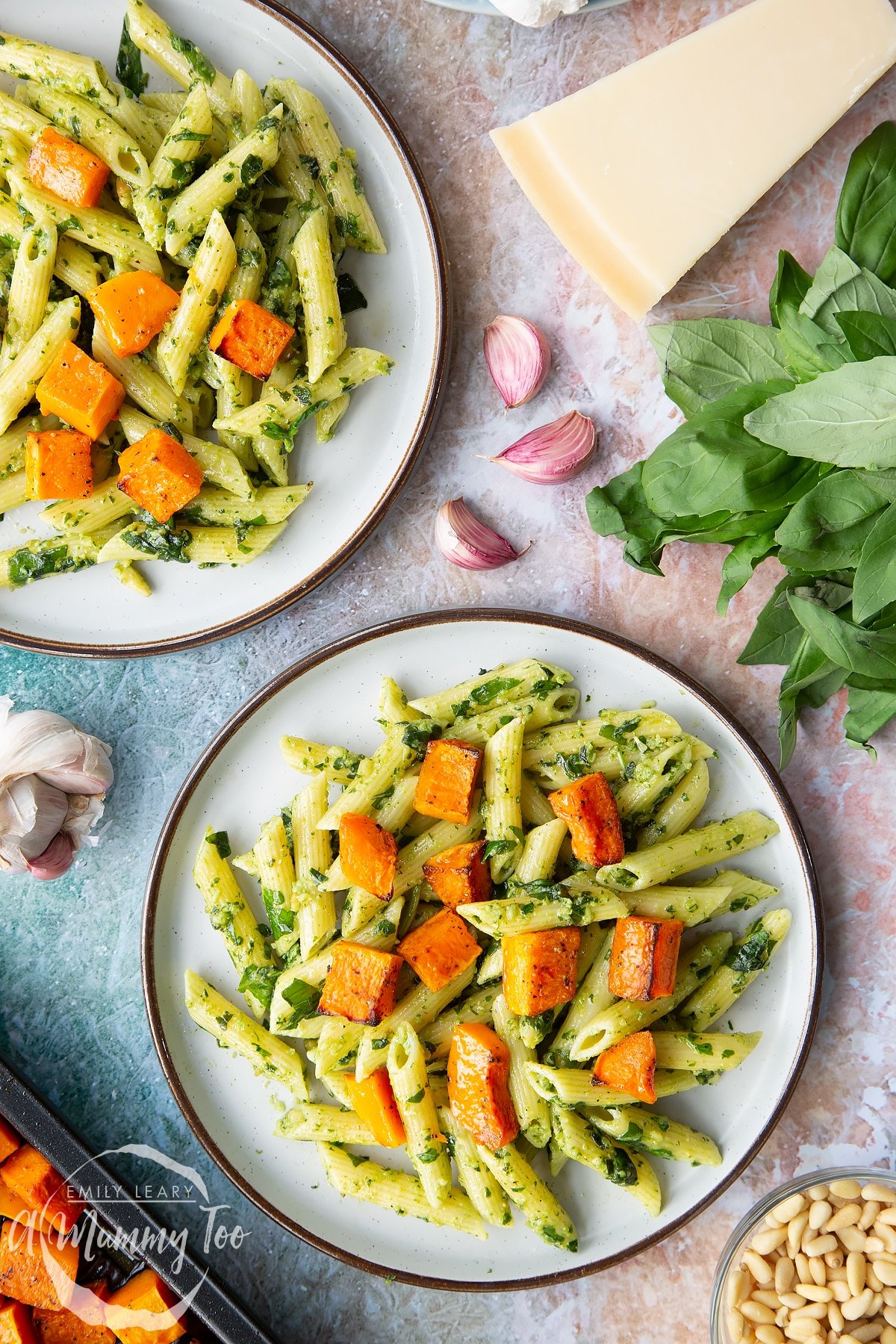 Penne pasta tossed with fresh pesto, served to plates and topped with roasted butternut squash. Ingredients to make penne with butternut squash and goat cheese surround the plates.