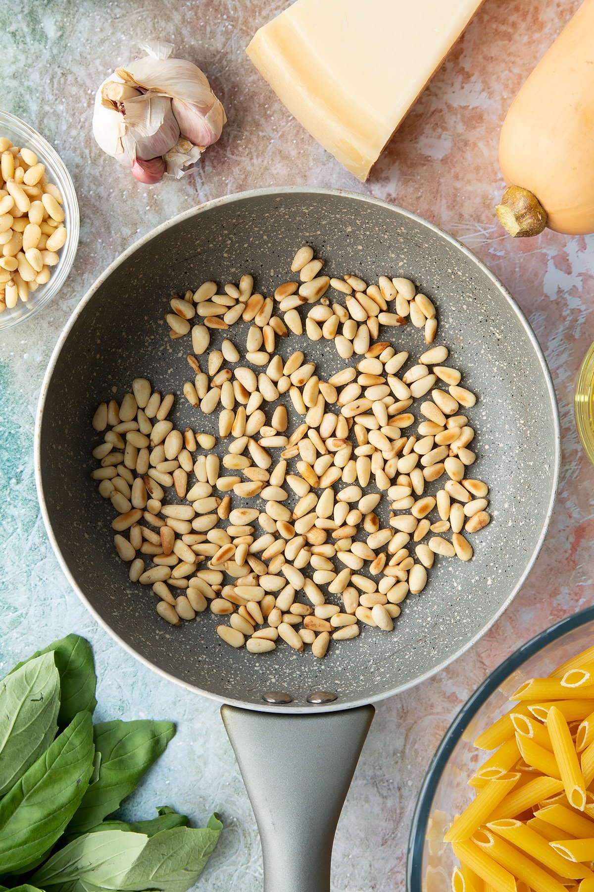 Toasted pine nuts in a small frying pan. Ingredients to make penne with butternut squash and goat cheese surround the pan.