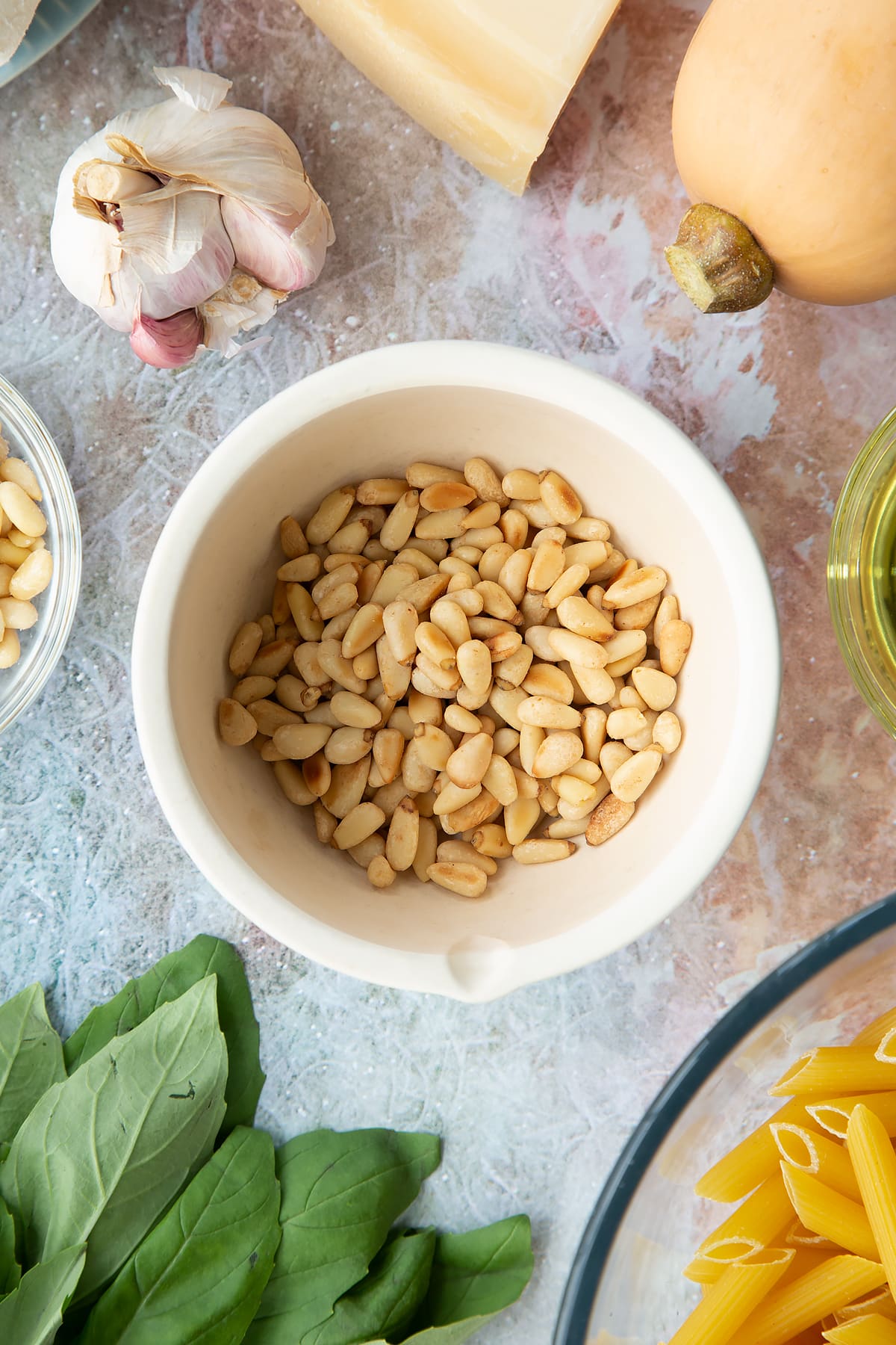 Toasted pine nuts in a small mortar. Ingredients to make penne with butternut squash and goat cheese surround the mortar.