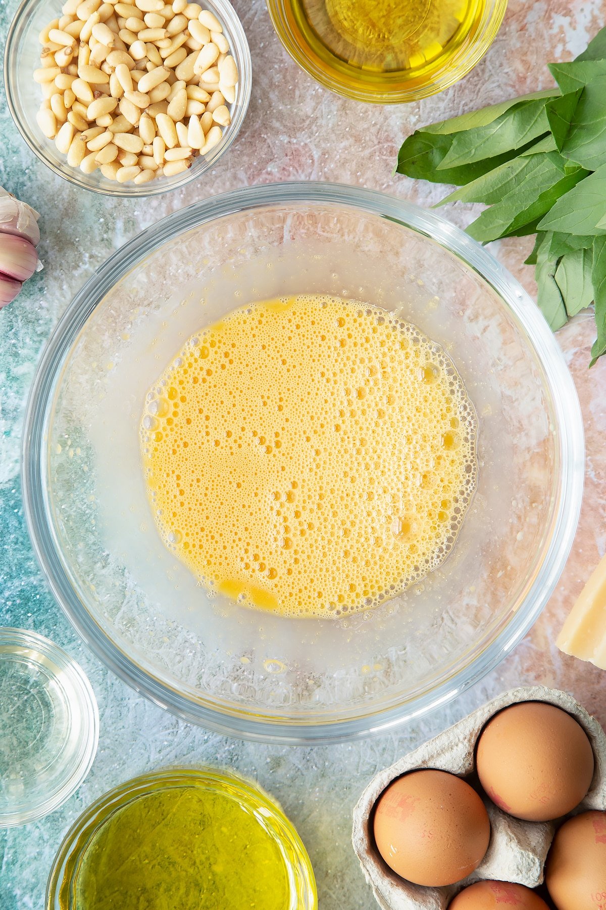 Egg yolks, mustard and vinegar whisked together in a bowl with oil on top. Ingredients to make pesto mayo surround the bowl.