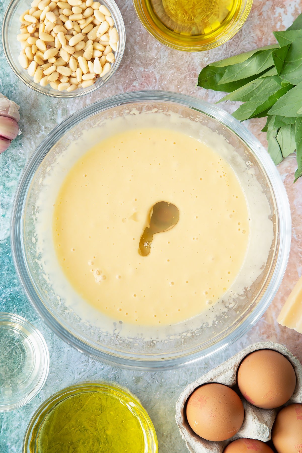 Egg yolks, mustard, vinegar and a little oil whisked together in a bowl with more oil on top. Ingredients to make pesto mayo surround the bowl.