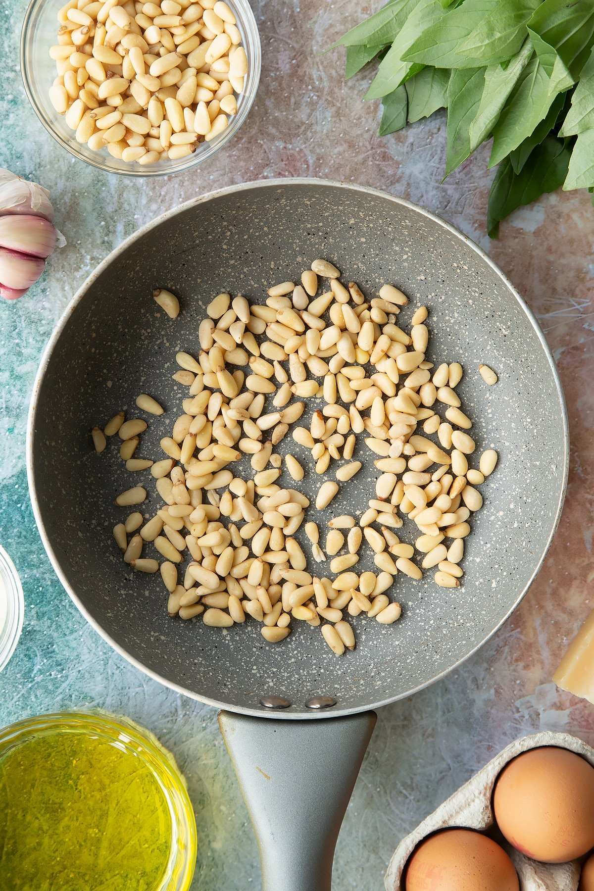 Pine nuts in a small frying pan. Ingredients to make pesto mayo surround the pan.