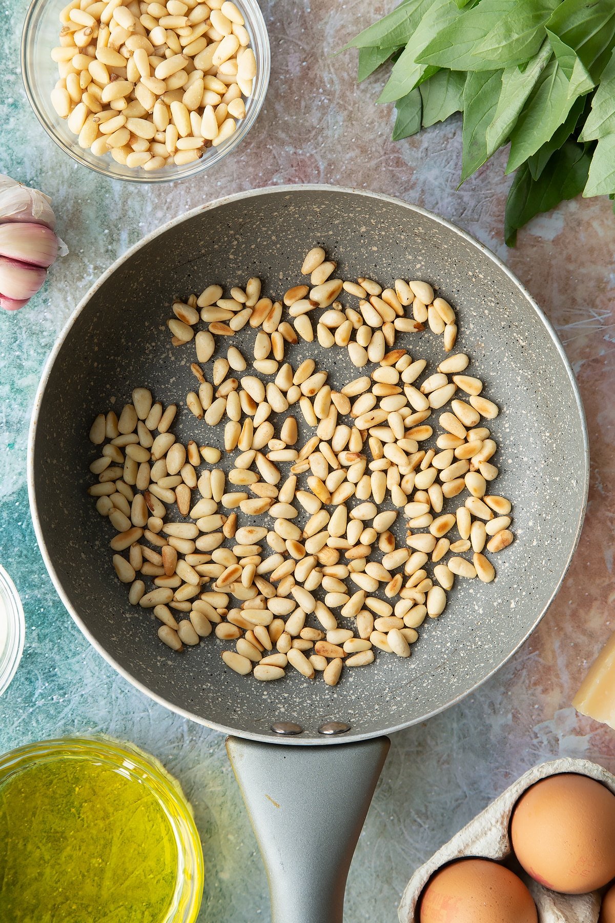 Toasted pine nuts in a small frying pan. Ingredients to make pesto mayo surround the pan.