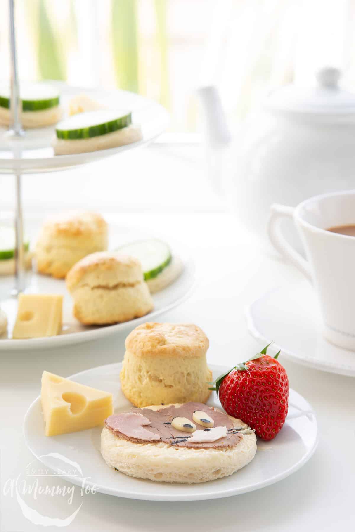 Forward facing shot of a plate made up of a Tom and Jerry sandwhich, scone, slice of cheese and a strawberry. In the background there's a cup of tea and a three tier cake stand with ingredients on.
