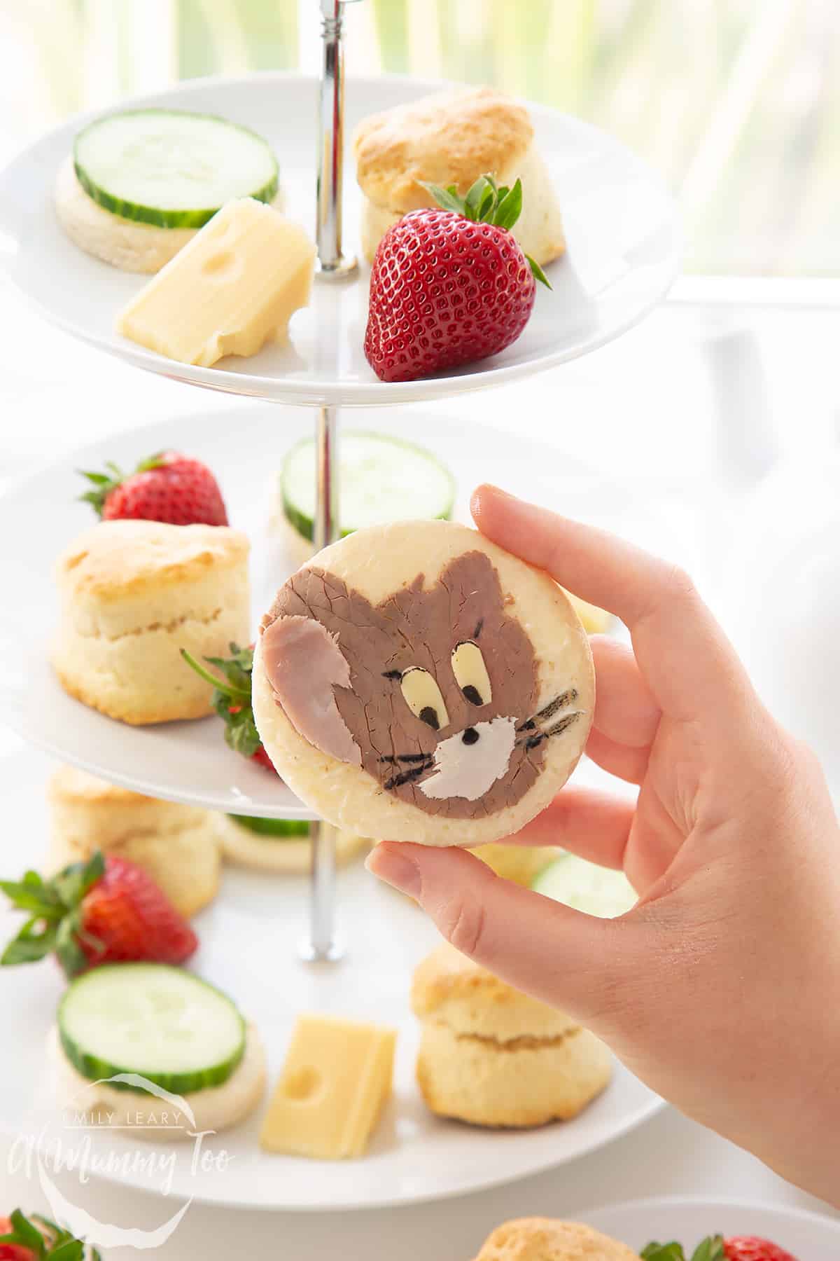 Hand holding up one of the Tom and Jerry sandwhiches with a cake stand filled with Tom and Jerry afternoon tea essentials in the background.