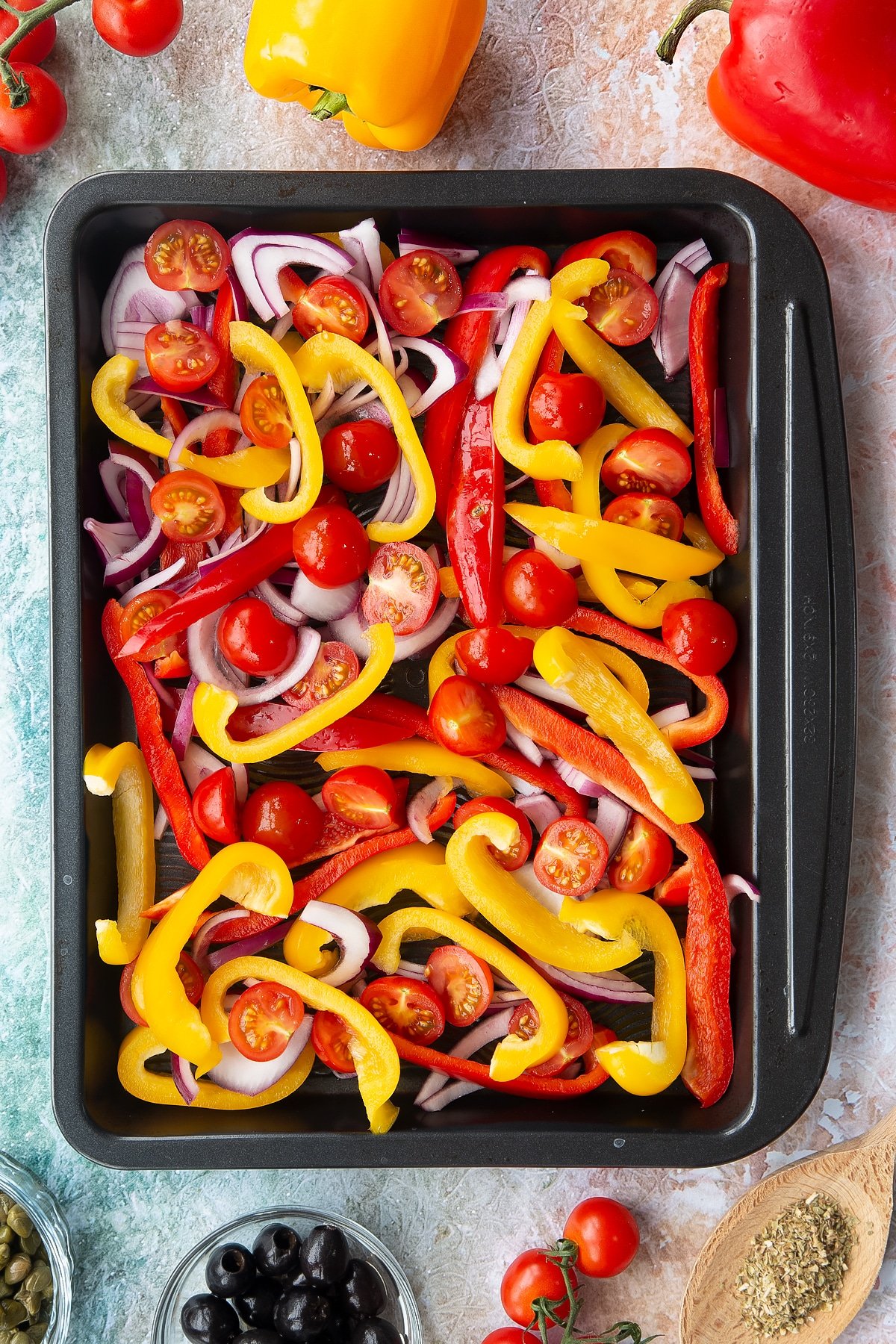 Halved cherry tomatoes, sliced chopped peppers and sliced onions in an oven tray. Ingredients to make vegan lentil salad surround the tray.