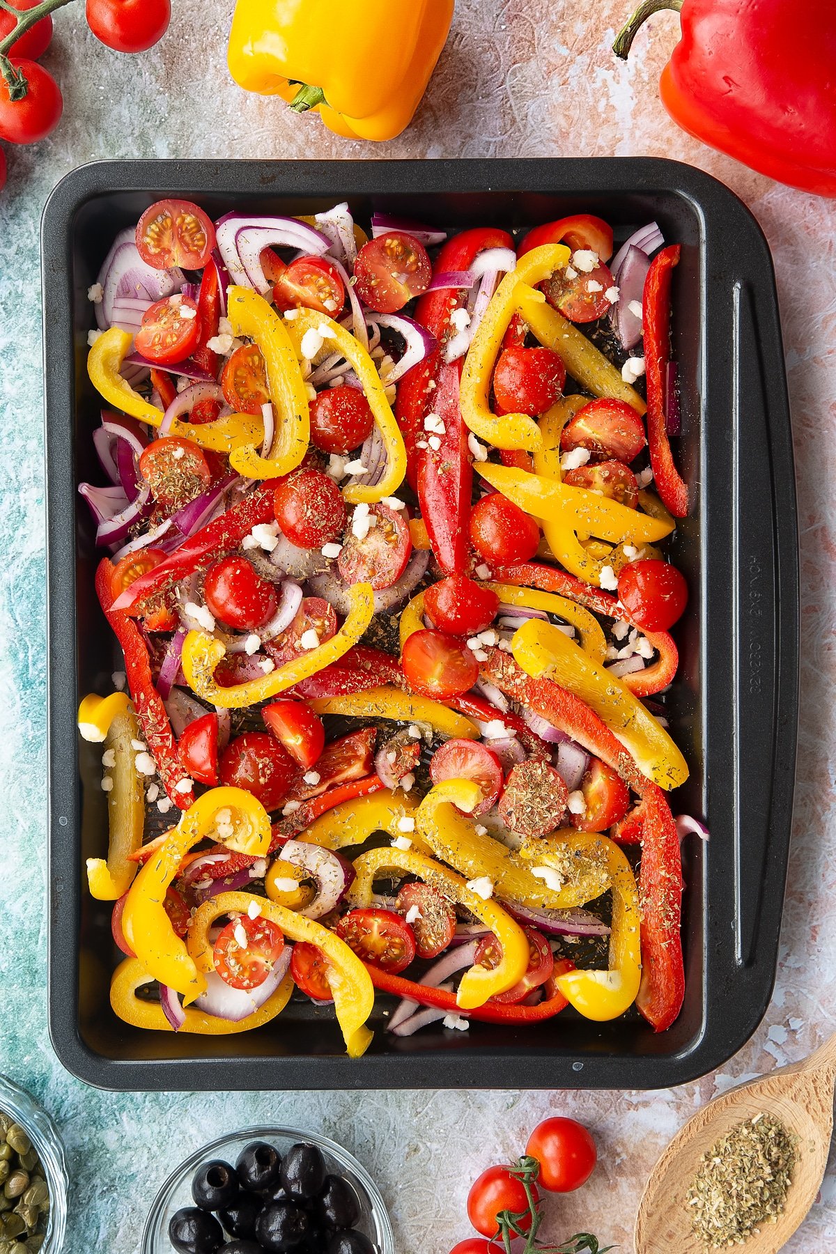 Halved cherry tomatoes, sliced chopped peppers and sliced onions with garlic, olive oil and oregano in an oven tray. Ingredients to make vegan lentil salad surround the tray.