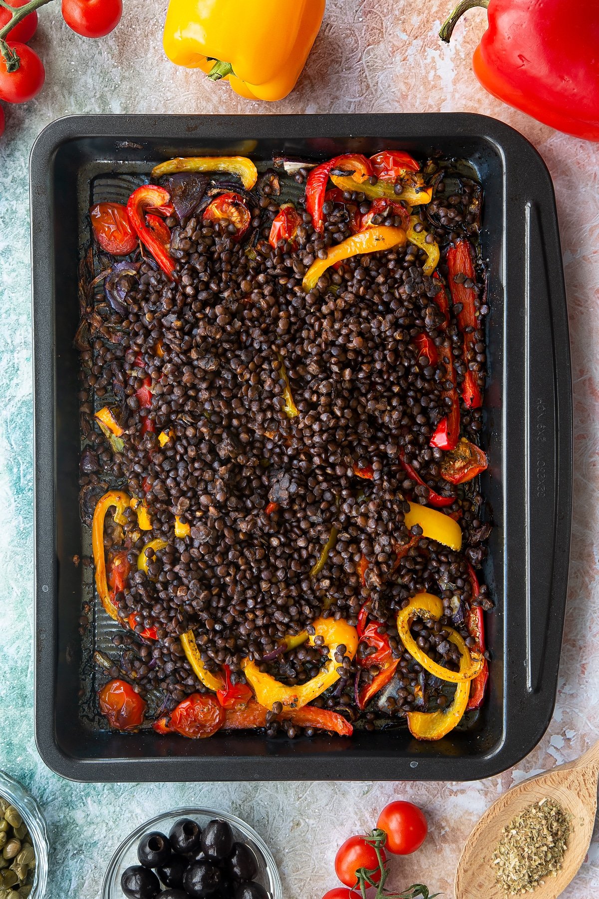 Roasted cherry tomatoes, peppers and onions in an oven tray, topped with cooked puy lentils. Ingredients to make vegan lentil salad surround the tray.